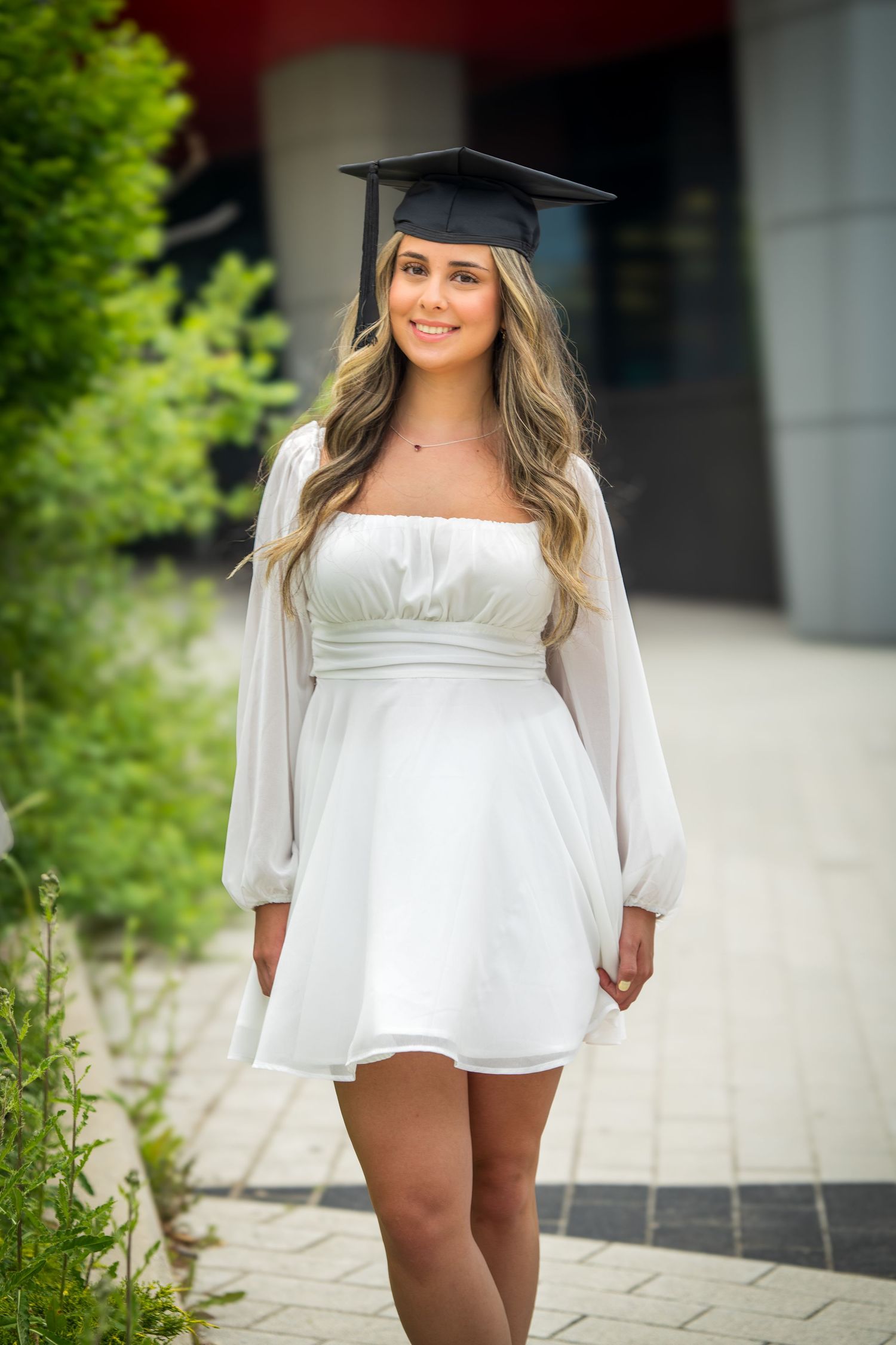 Graduate poses in white off-shoulder dress and cap on campus grounds.