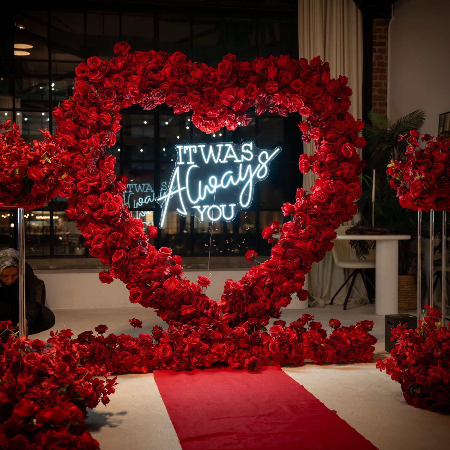 A romantic walkway decorated with red rose arches and illuminated neon signs reading 'It was Always'.