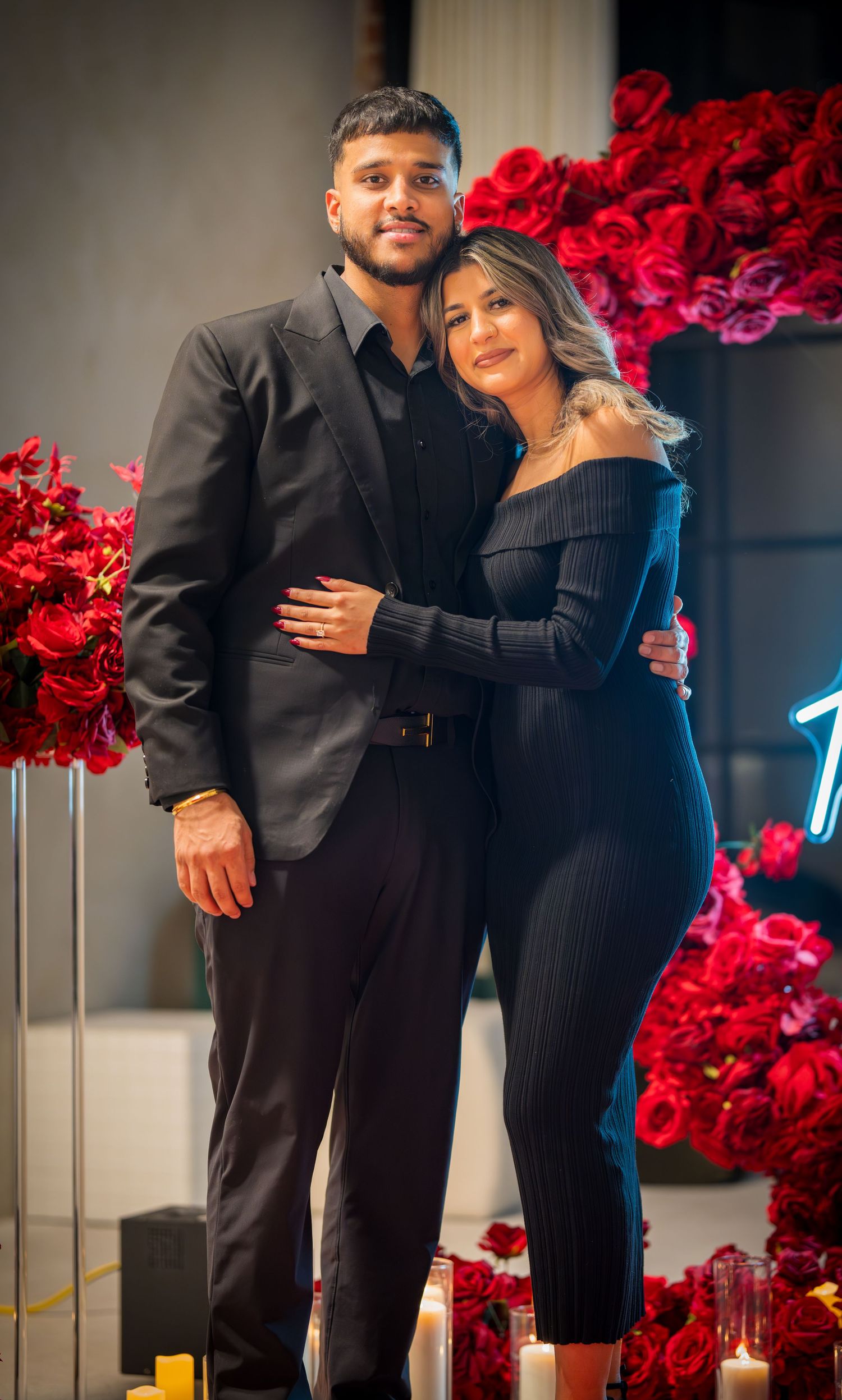 Romantic couple embraces in matching black outfits in front of a wall decorated with vibrant red roses.