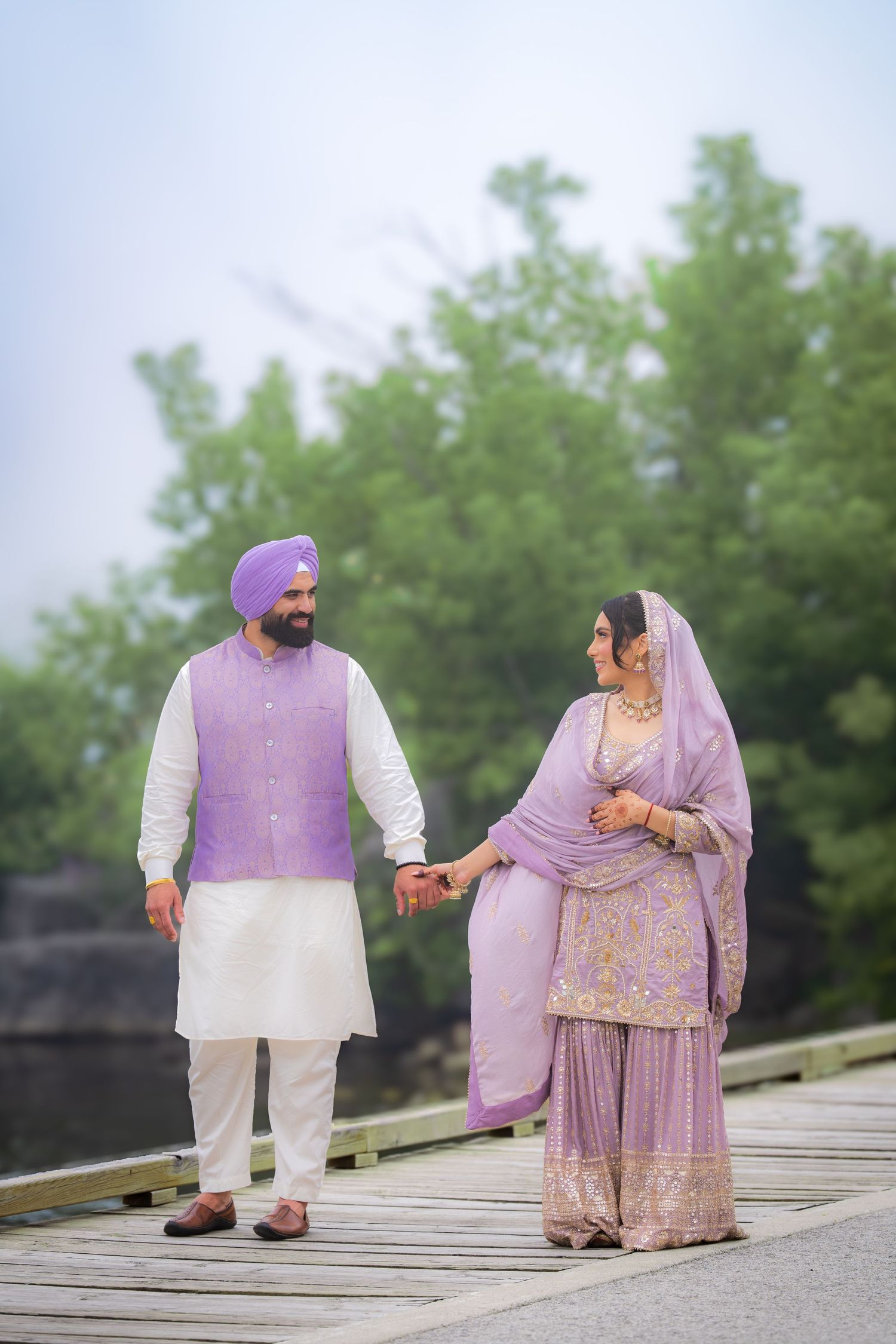 A couple in coordinated purple attire poses on a wooden deck surrounded by greenery.