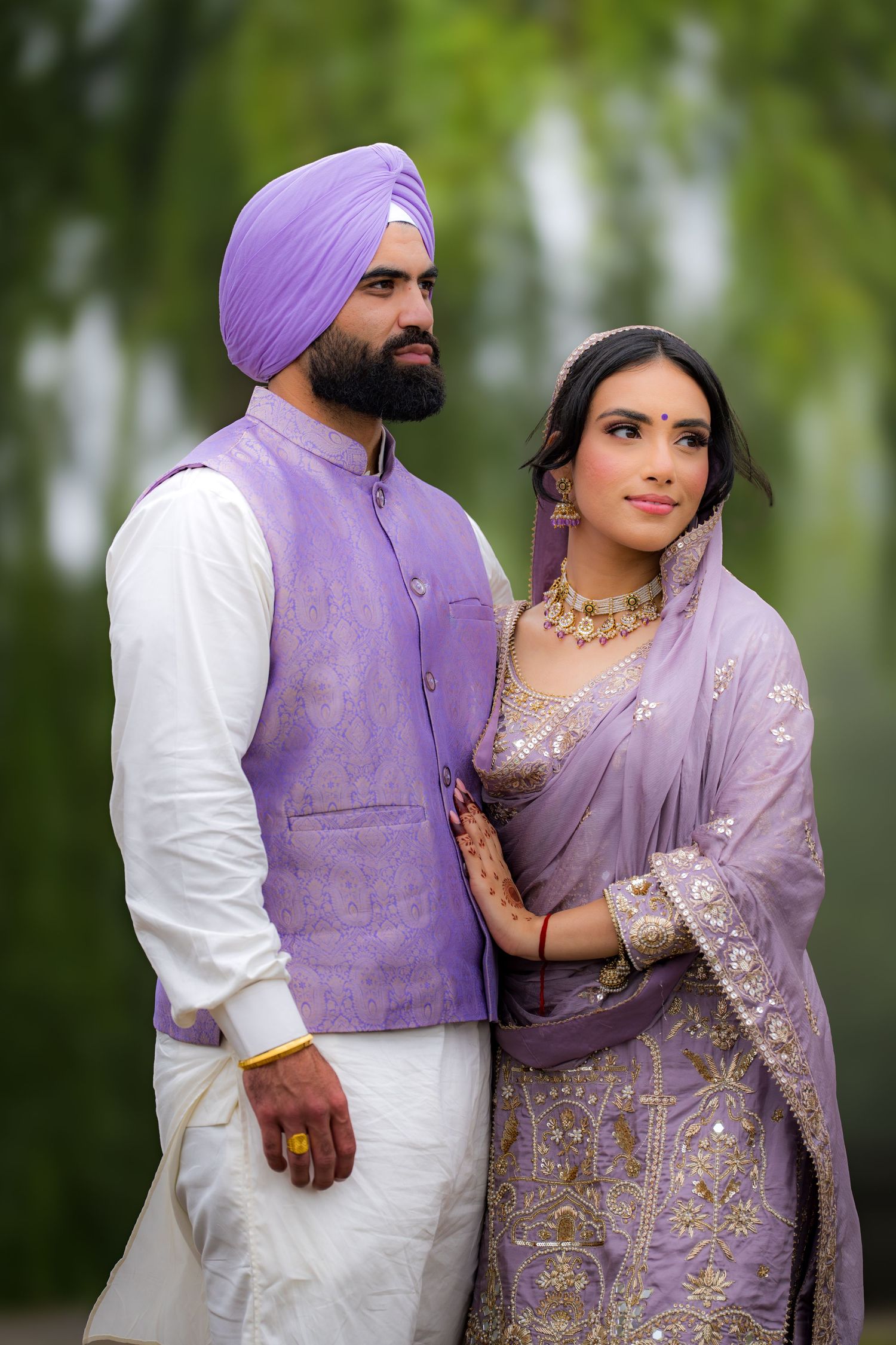 A couple in elegant Punjabi attire poses intimately together under willow trees wearing purple and white.