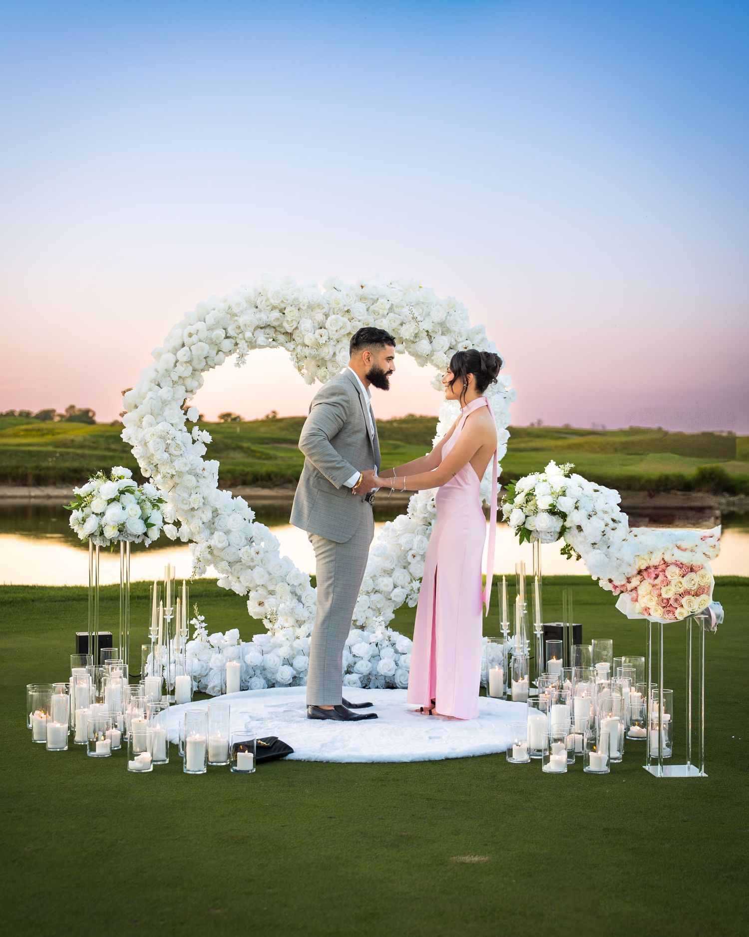 Elegant outdoor wedding setup with white floral arches and glass candle holders at sunset.