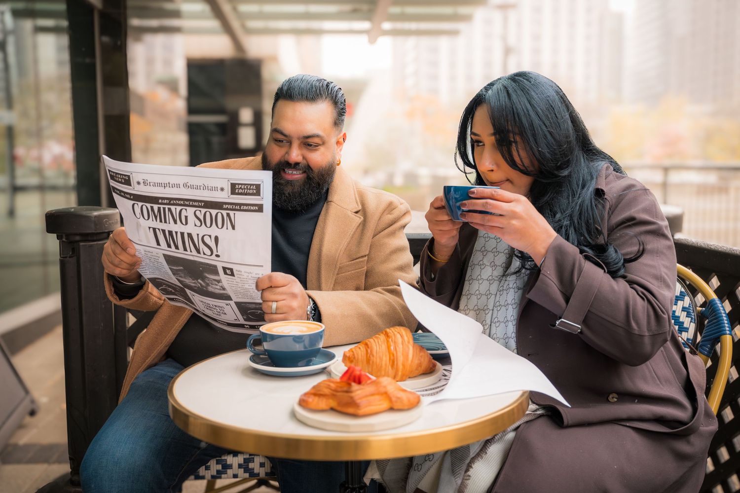 Couple enjoying breakfast at outdoor cafe with newspaper and pastries on a round table.