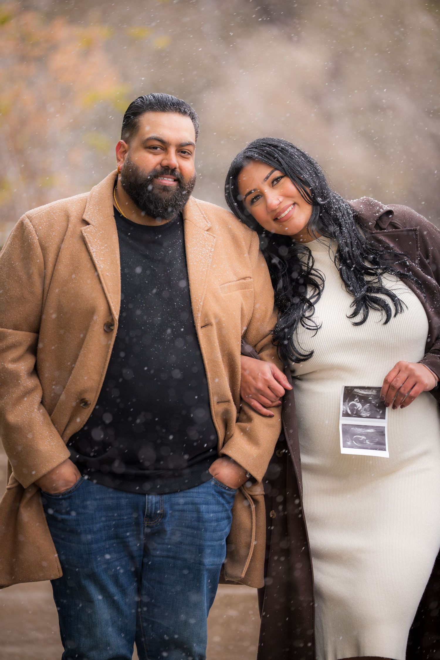A couple holds up a sonogram photo during a snowy winter pregnancy announcement photoshoot.