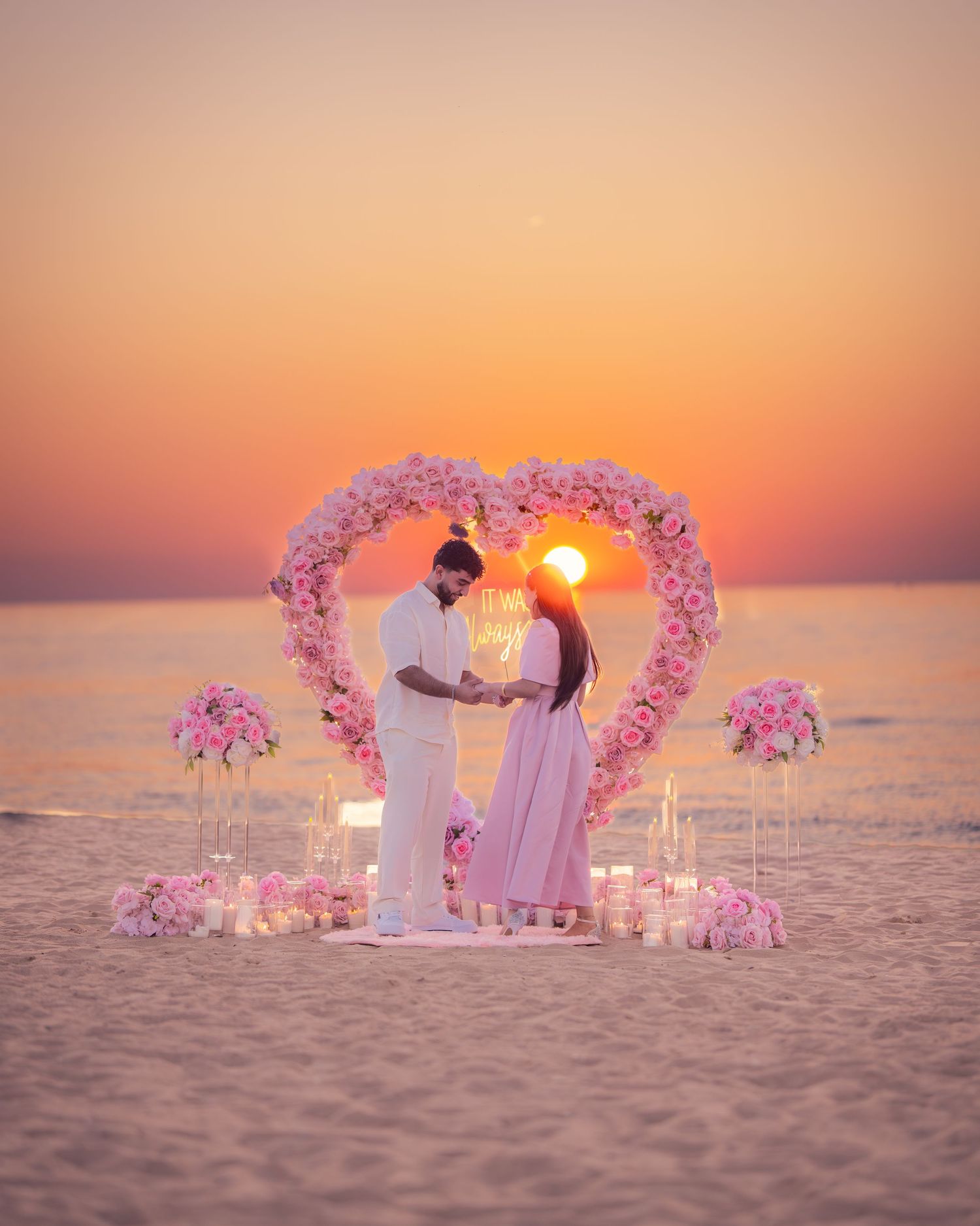 Beachfront marriage proposal scene with pink floral heart arch at sunset.