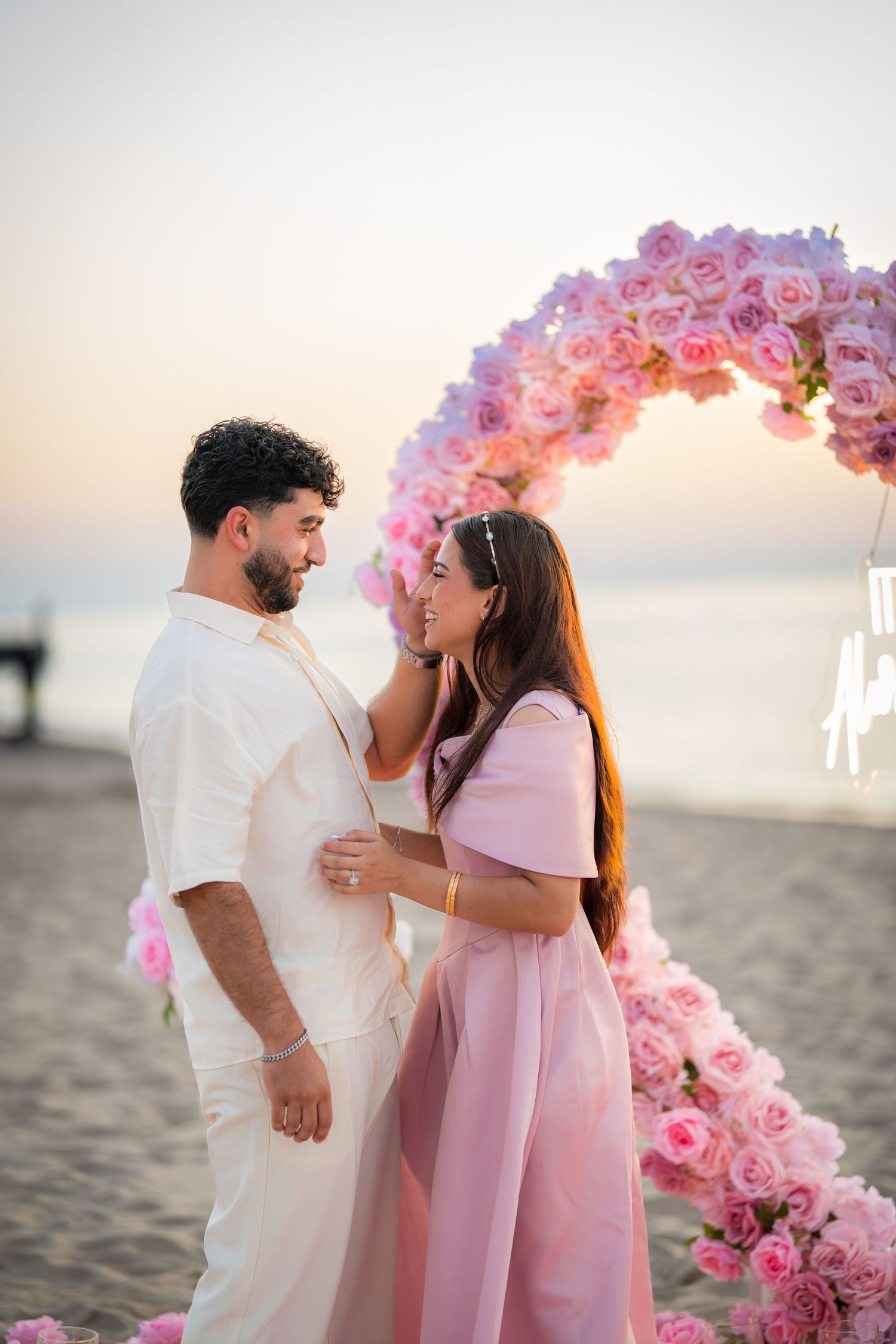 Romantic beach proposal scene with pink floral arch and sunset backdrop as couple shares intimate moment.