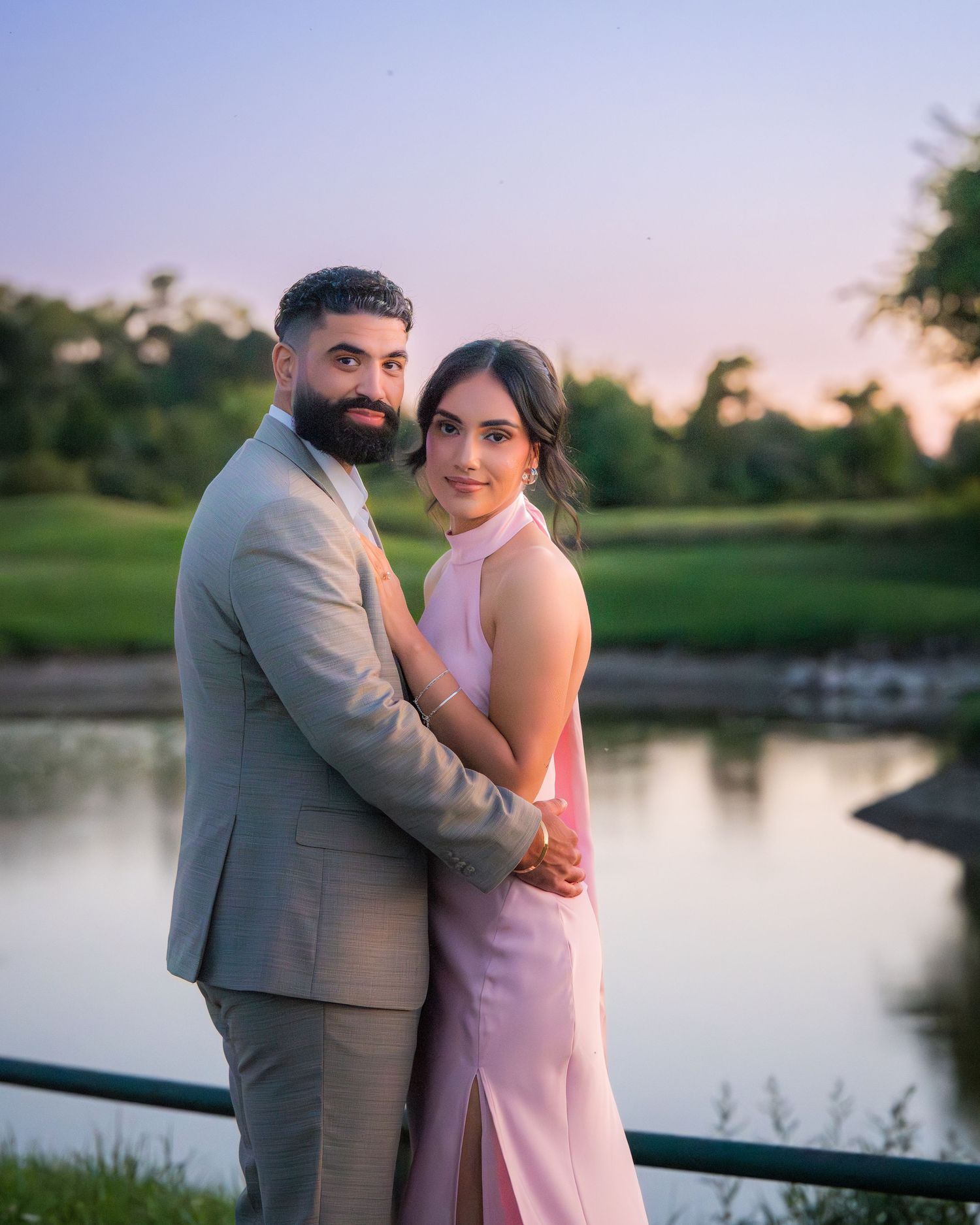 A couple embraces by a tranquil lake at sunset wearing formal pink and grey outfits.