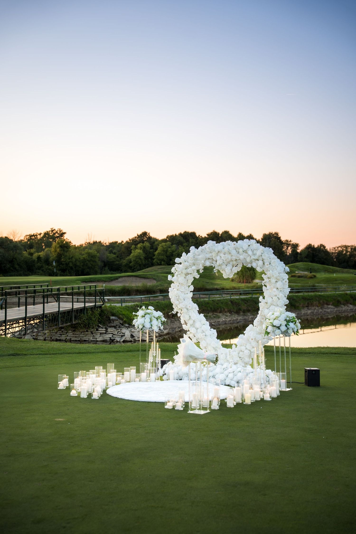 Outdoor proposal setting with curved white floral arch and lanterns on green grass.