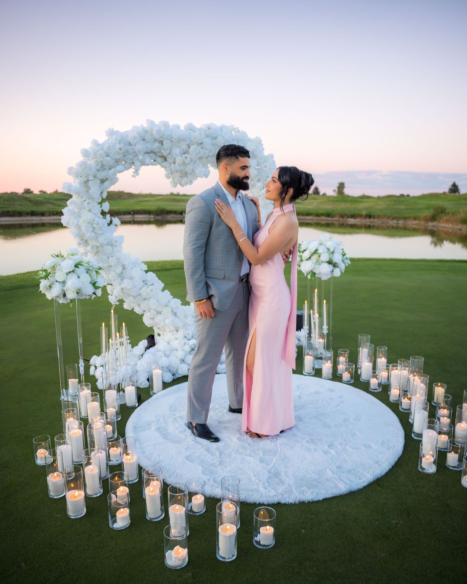 Romantic wedding scene on golf course with white floral arch and lantern-lined aisle at dusk.