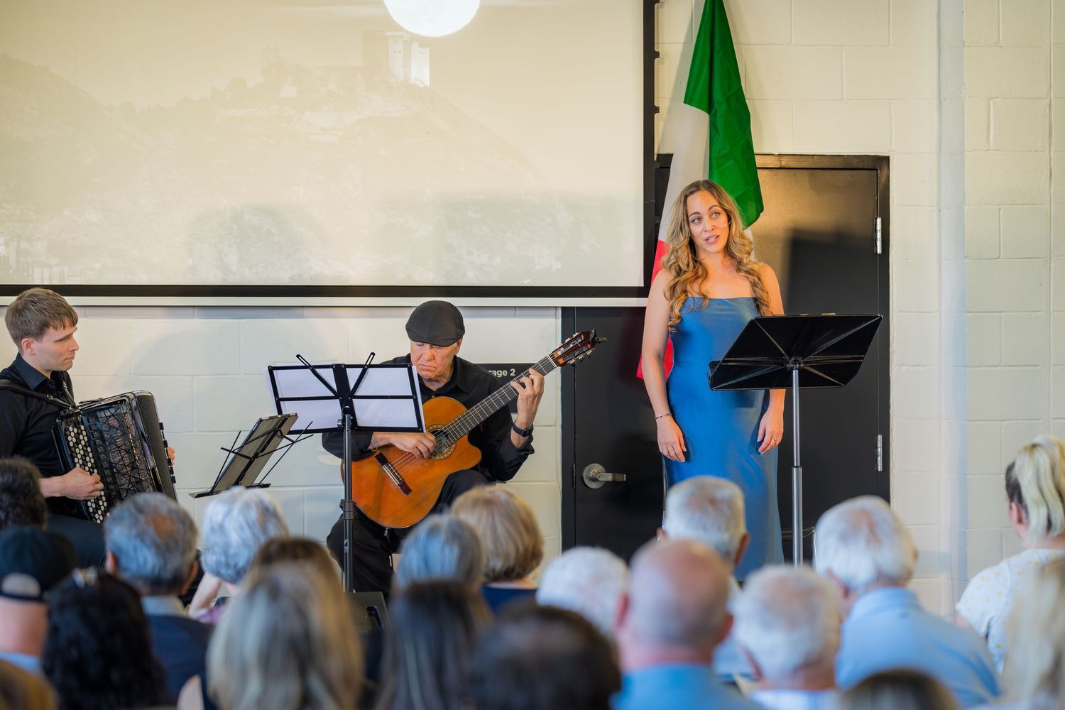 Trio of musicians performing live with accordion, guitar, and soprano vocalist for seated audience.