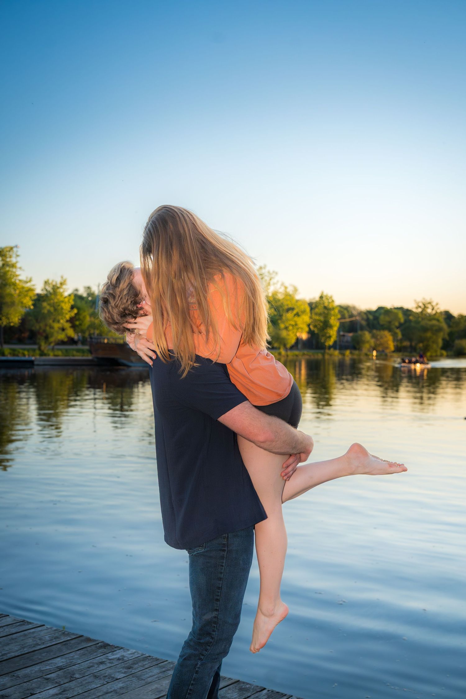 Man lifting woman in his arms on a wooden dock at sunset with a tranquil lake behind them.