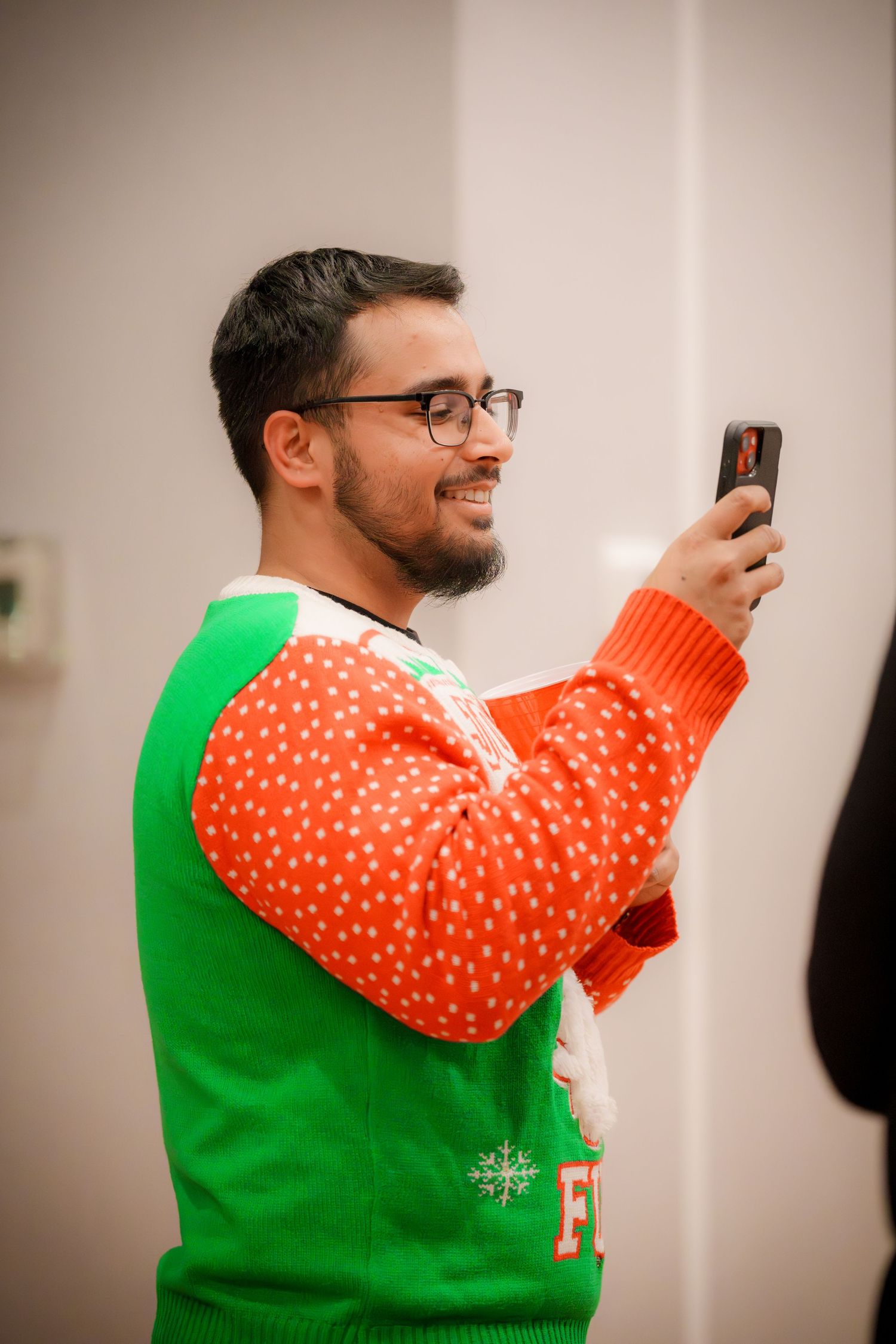 Person in festive orange and green Christmas sweater smiling while using phone at party.