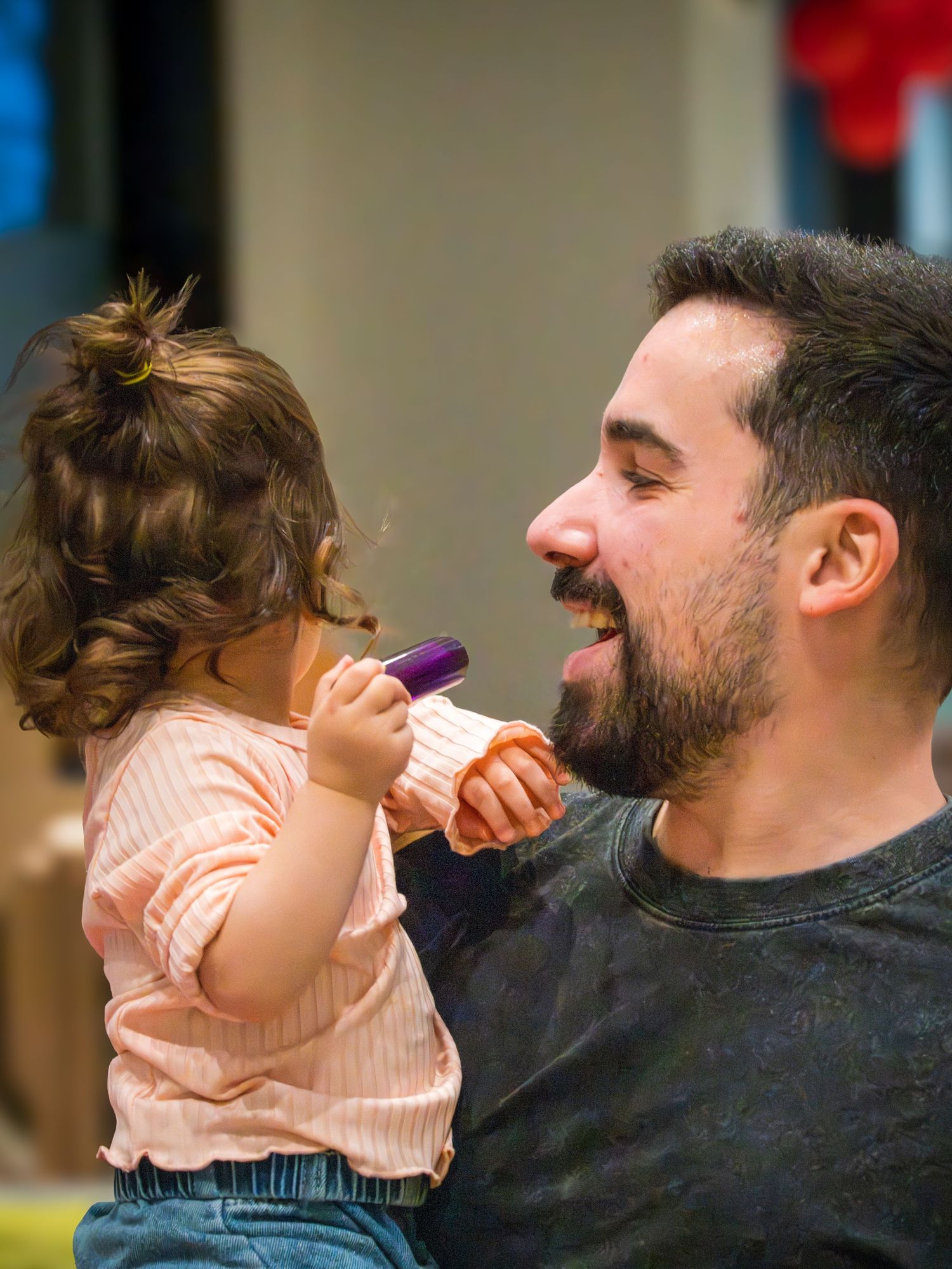 A bearded man playfully pretends to eat a purple popsicle held by a toddler girl in a pink top.