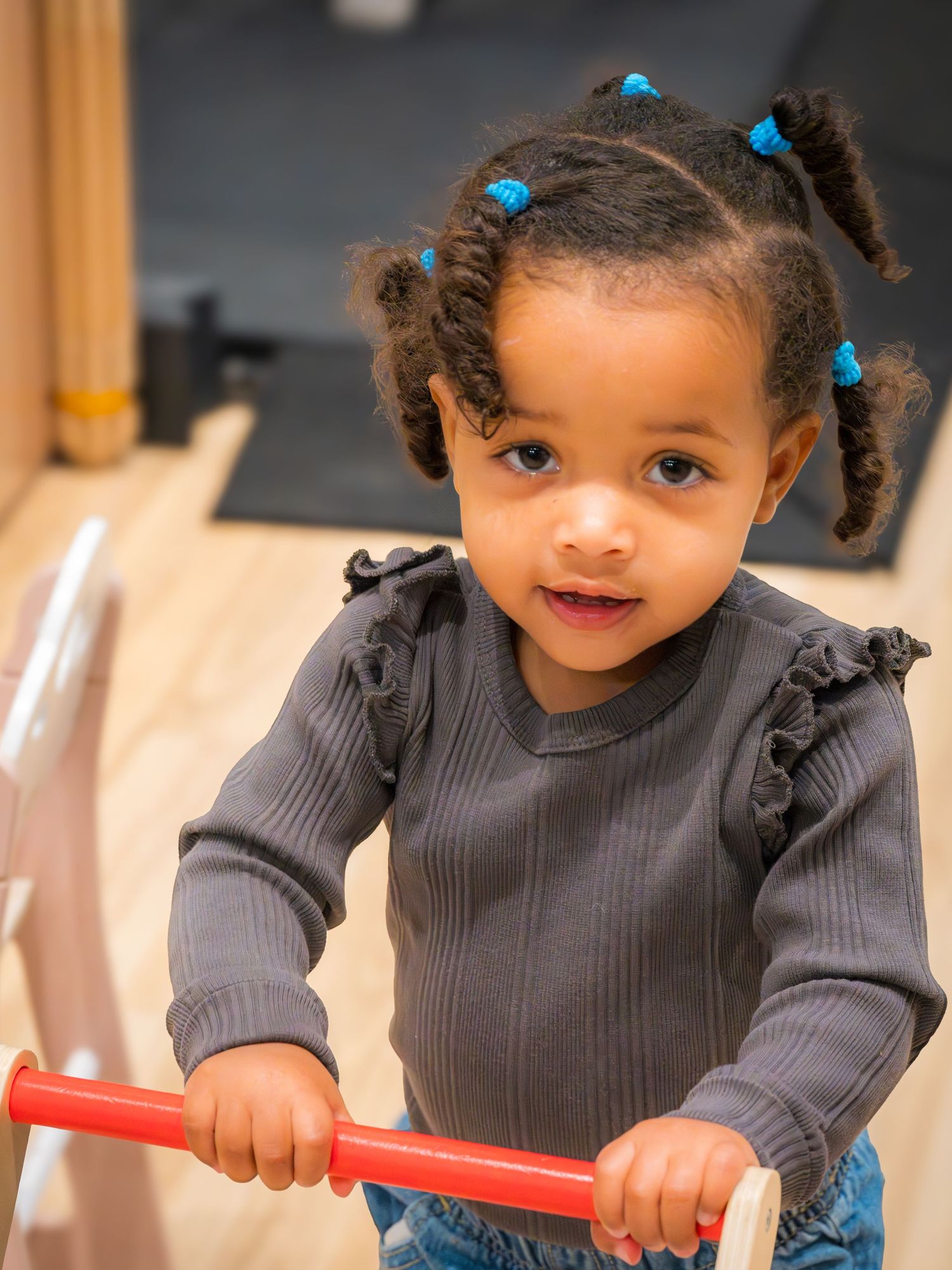 Cute toddler girl with blue hair ties riding a wooden balance bike indoors.