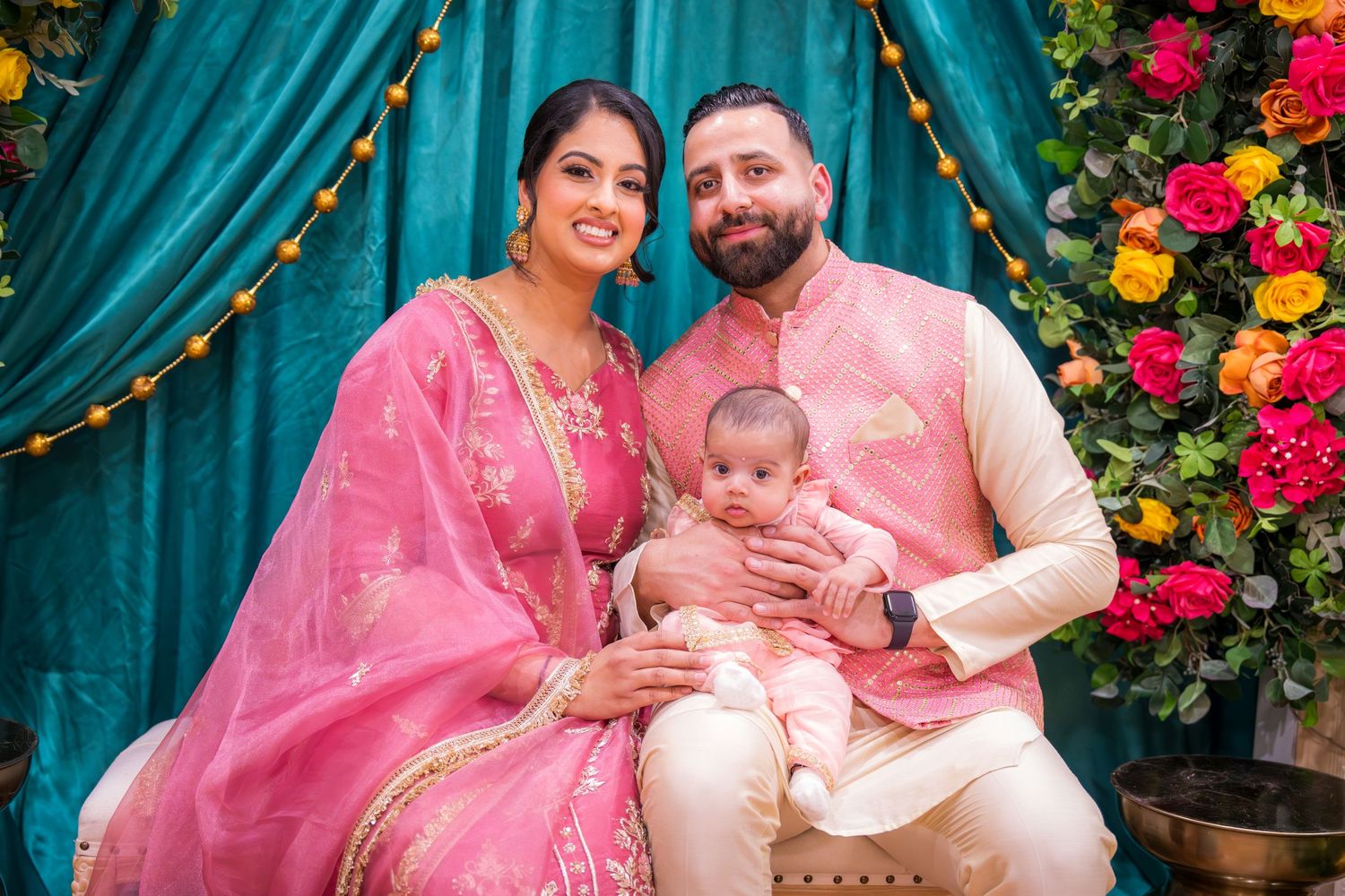 South Asian family with newborn baby posing before teal and floral ceremony backdrop.
