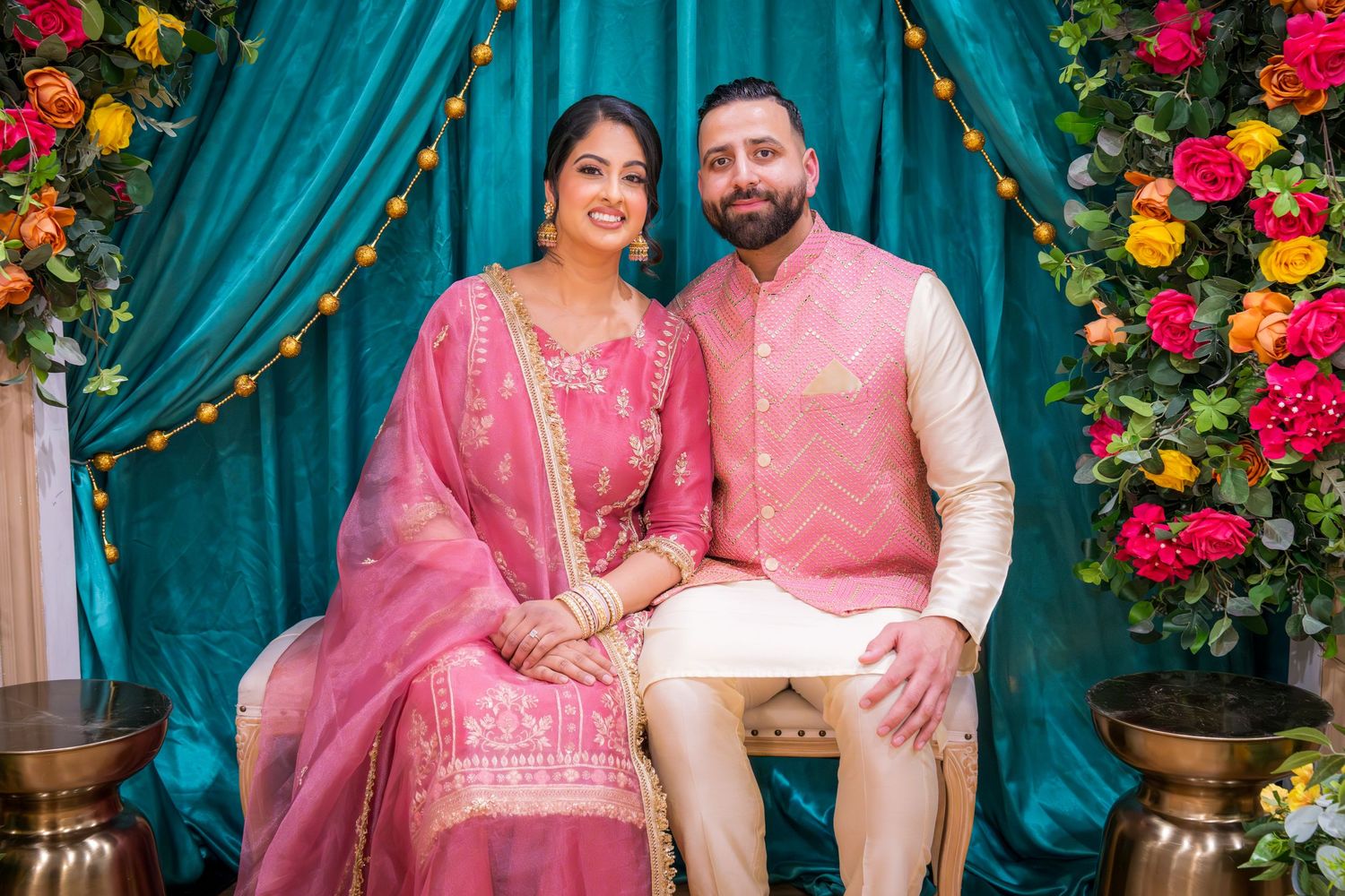 Indian couple in coordinating pink and cream traditional outfits seated together at vibrant floral decorated ceremony.