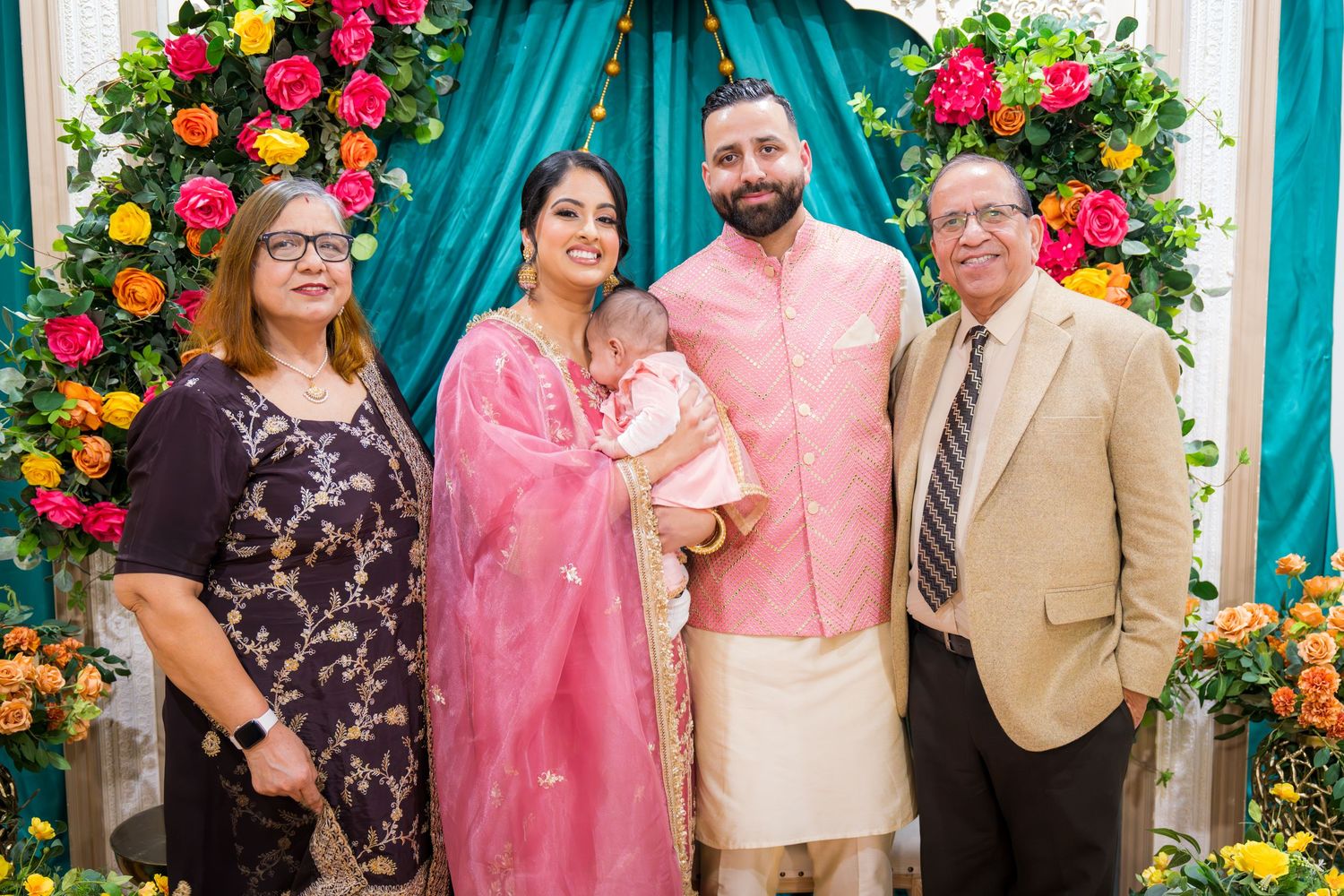 Four adults holding newborn baby posing before vibrant floral arch at Indian celebration.