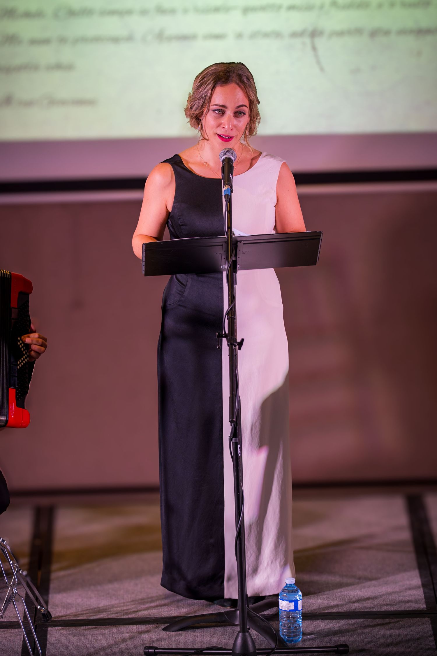 Elegant woman in two-tone black and white gown reading at a music stand microphone during a formal event.