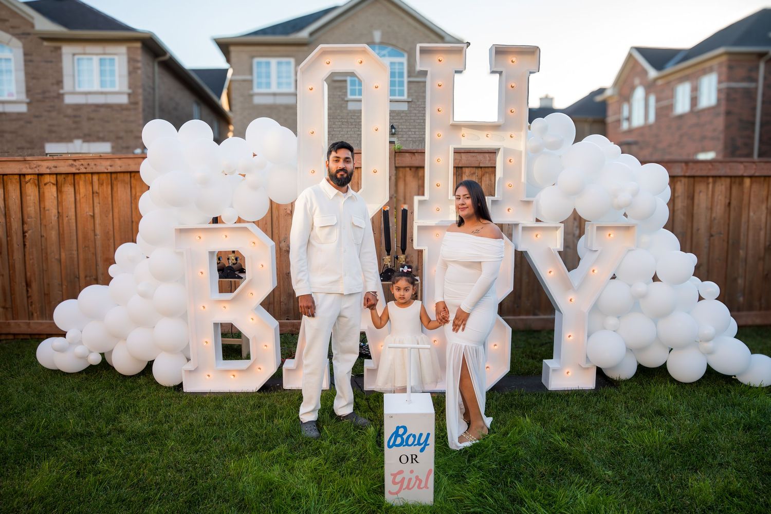 Family of three poses in front of OH BABY marquee letters with white balloons at gender reveal party.