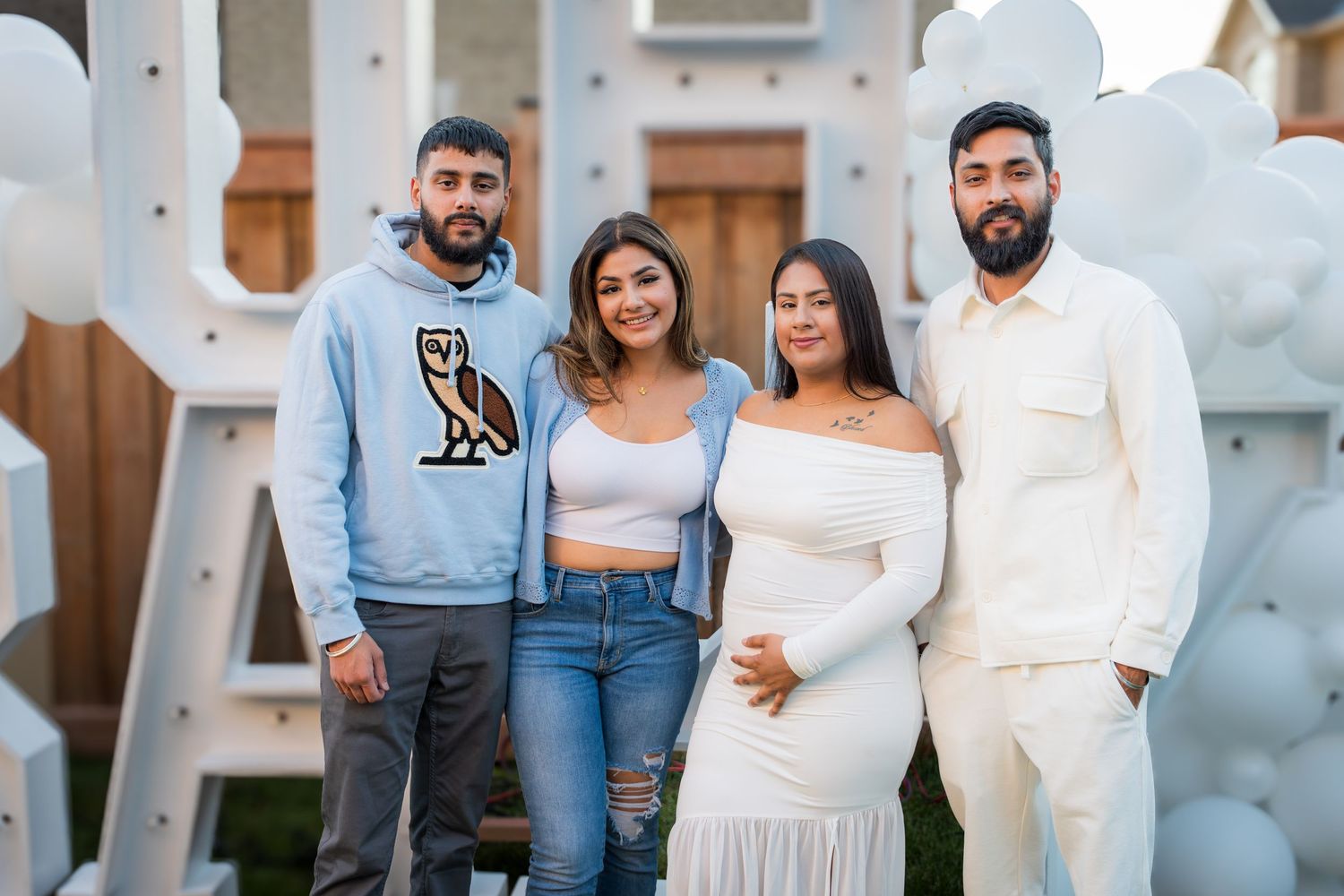 Four people pose together at a baby shower with white balloon decorations and large letter props.