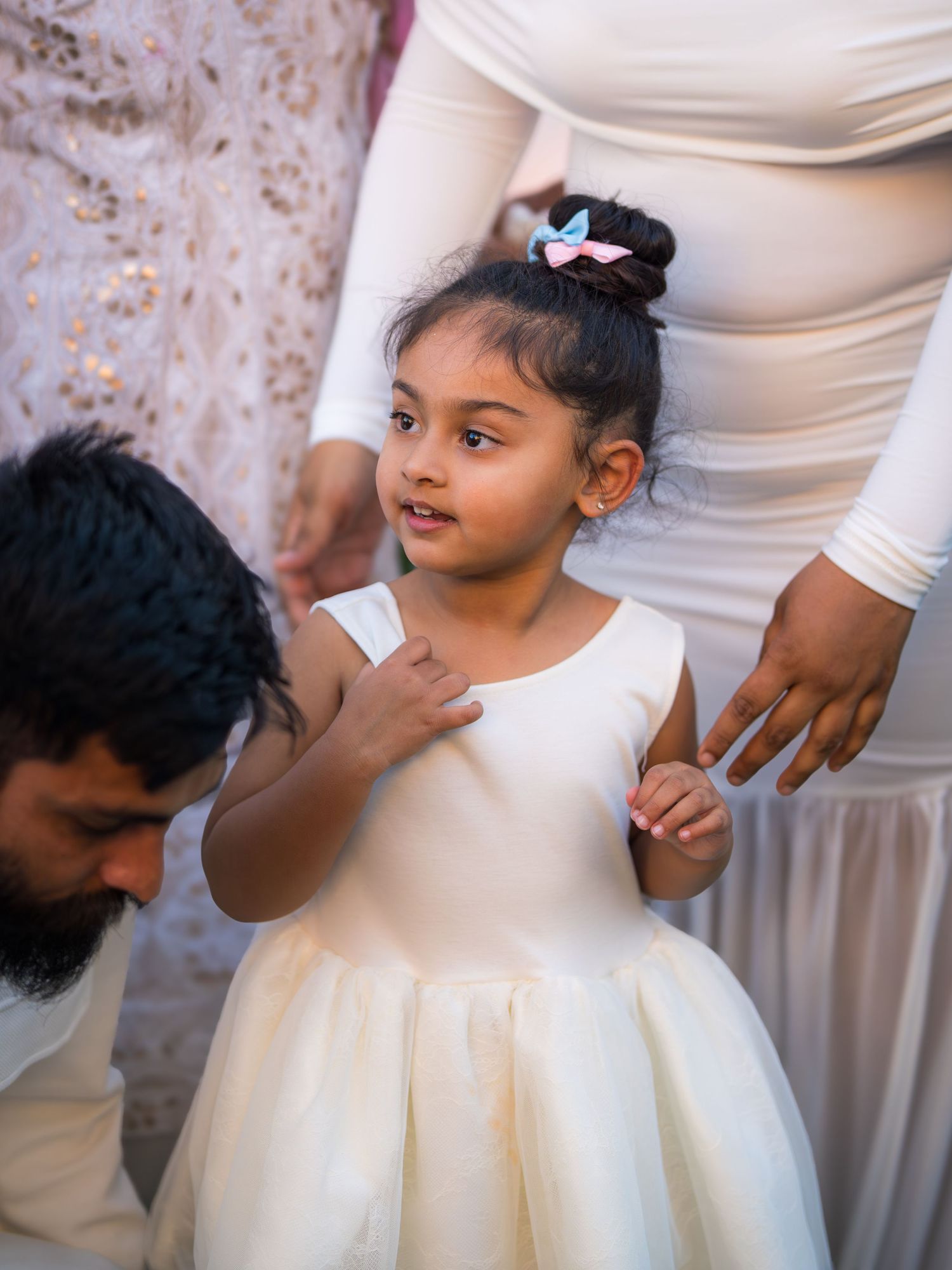 Young girl in white dress at a baby shower standing between parents with a bow in her hair.