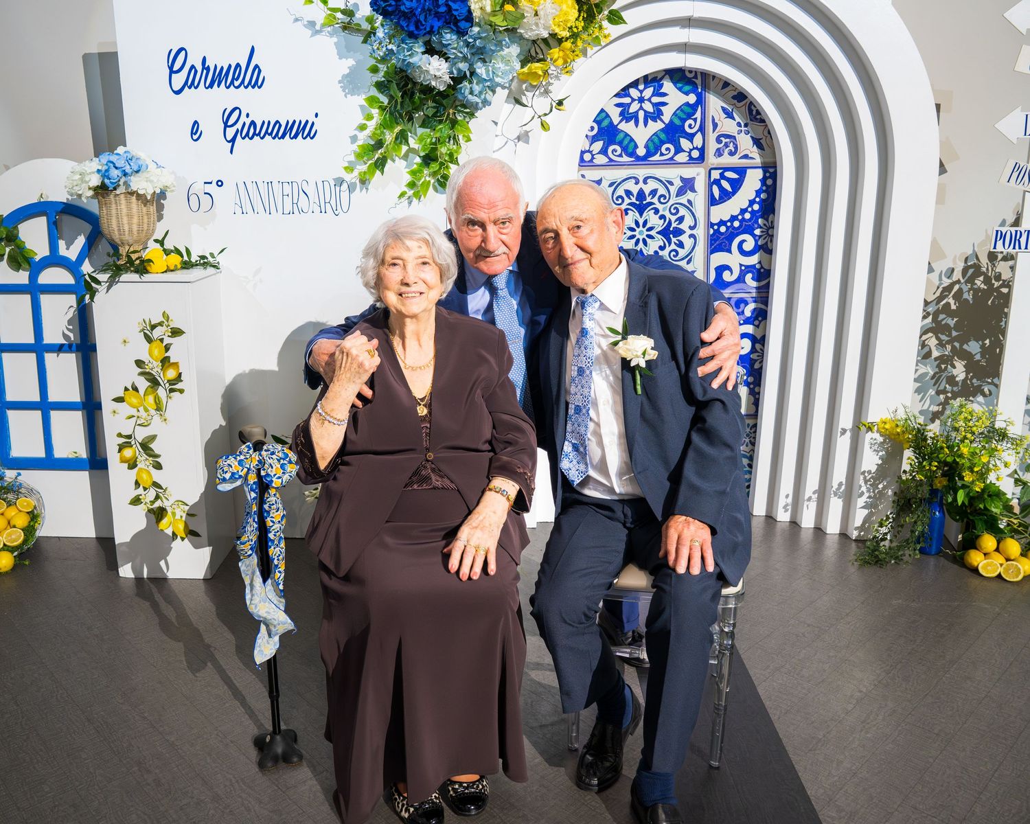 Elderly anniversary couple seated with male guest in front of Carmela and Giovanni 65th anniversary floral backdrop.