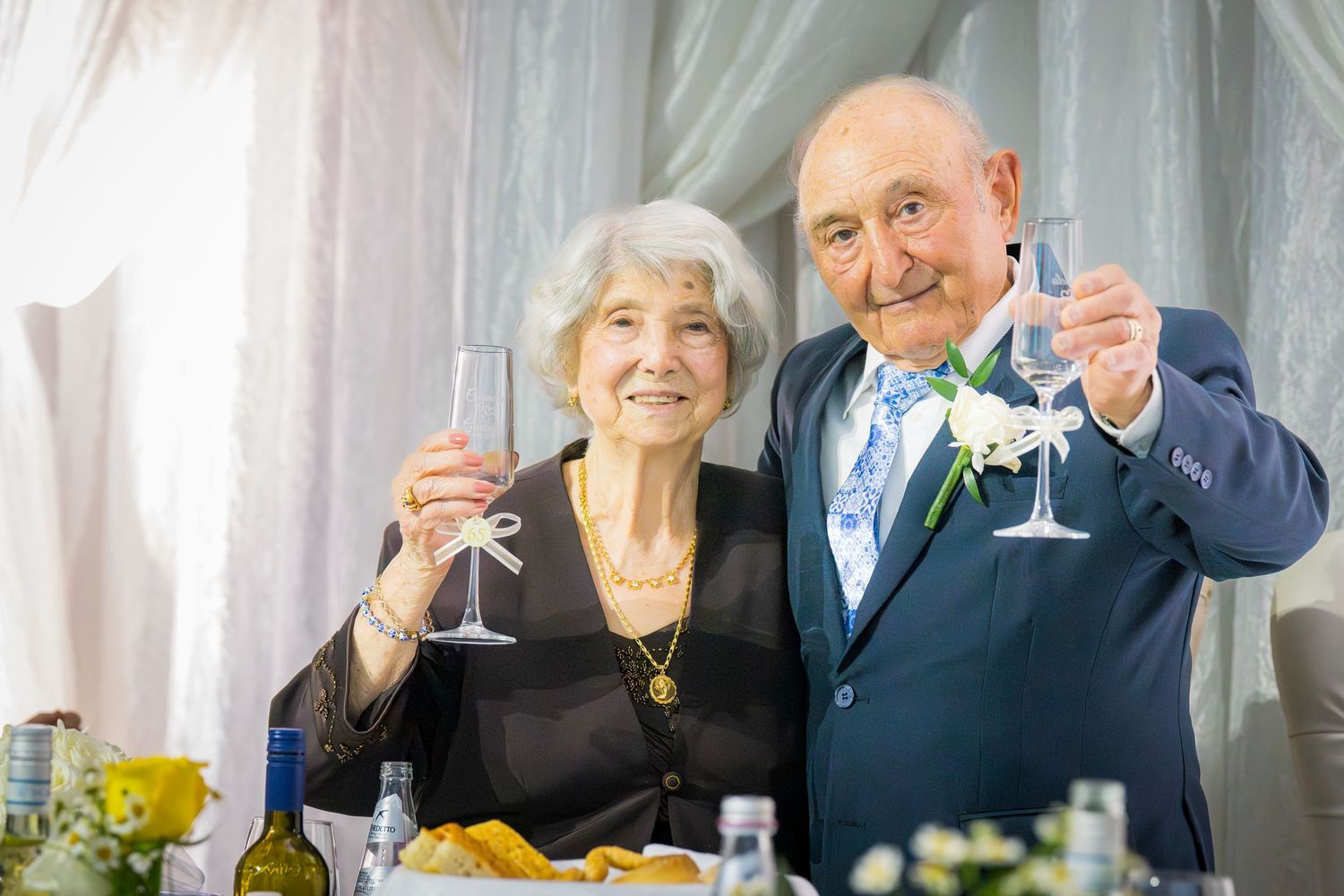 Senior couple toasting with champagne glasses at anniversary celebration, man in blue suit with floral boutonniere.