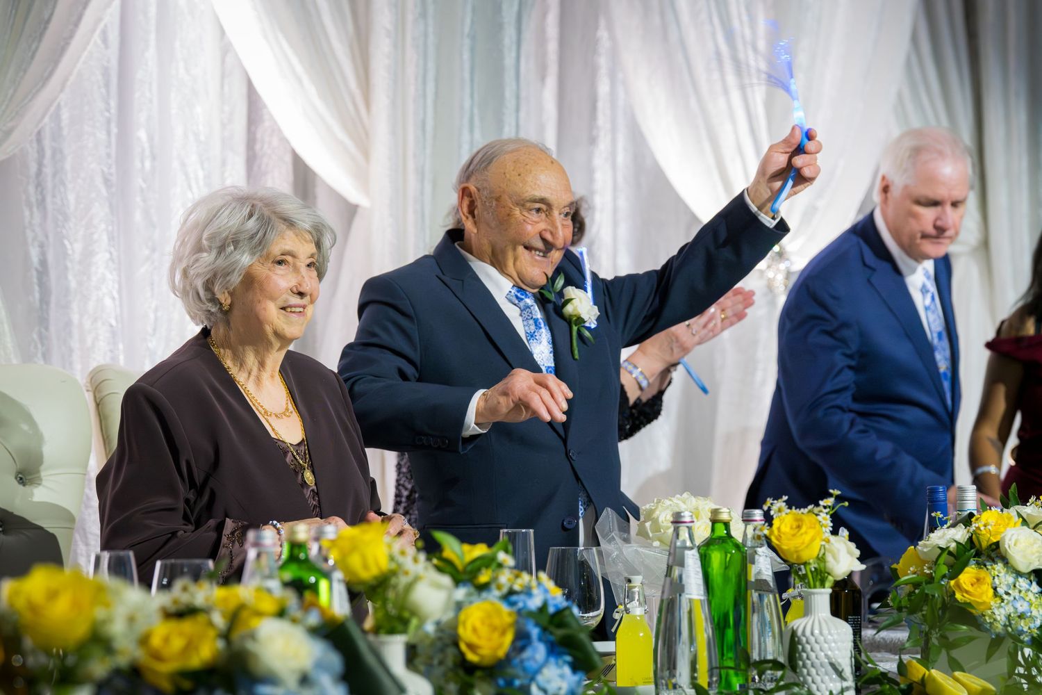 Elderly couple celebrating at a formal event, man raising a glass in a toast surrounded by yellow floral arrangements.