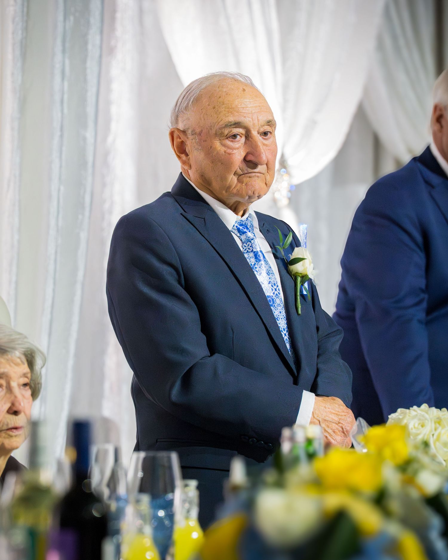 Elderly man in navy suit with floral tie and boutonniere standing at an anniversary celebration dinner table.