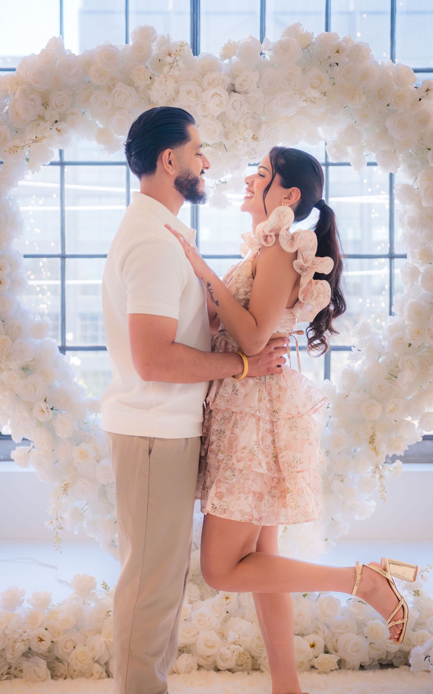 Couple embracing and smiling under a white floral heart arch during a romantic proposal setup.