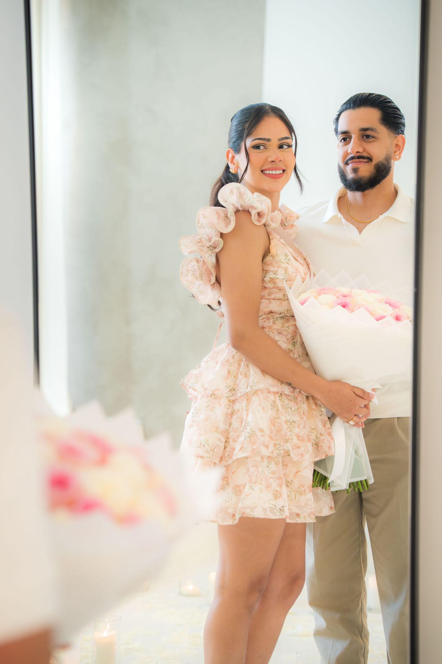 Couple posing together in mirror reflection, woman in floral dress holding pink rose bouquet.
