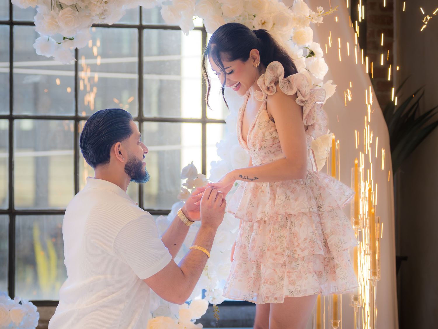 Man proposing to woman in floral dress with white flower arch and sparklers.