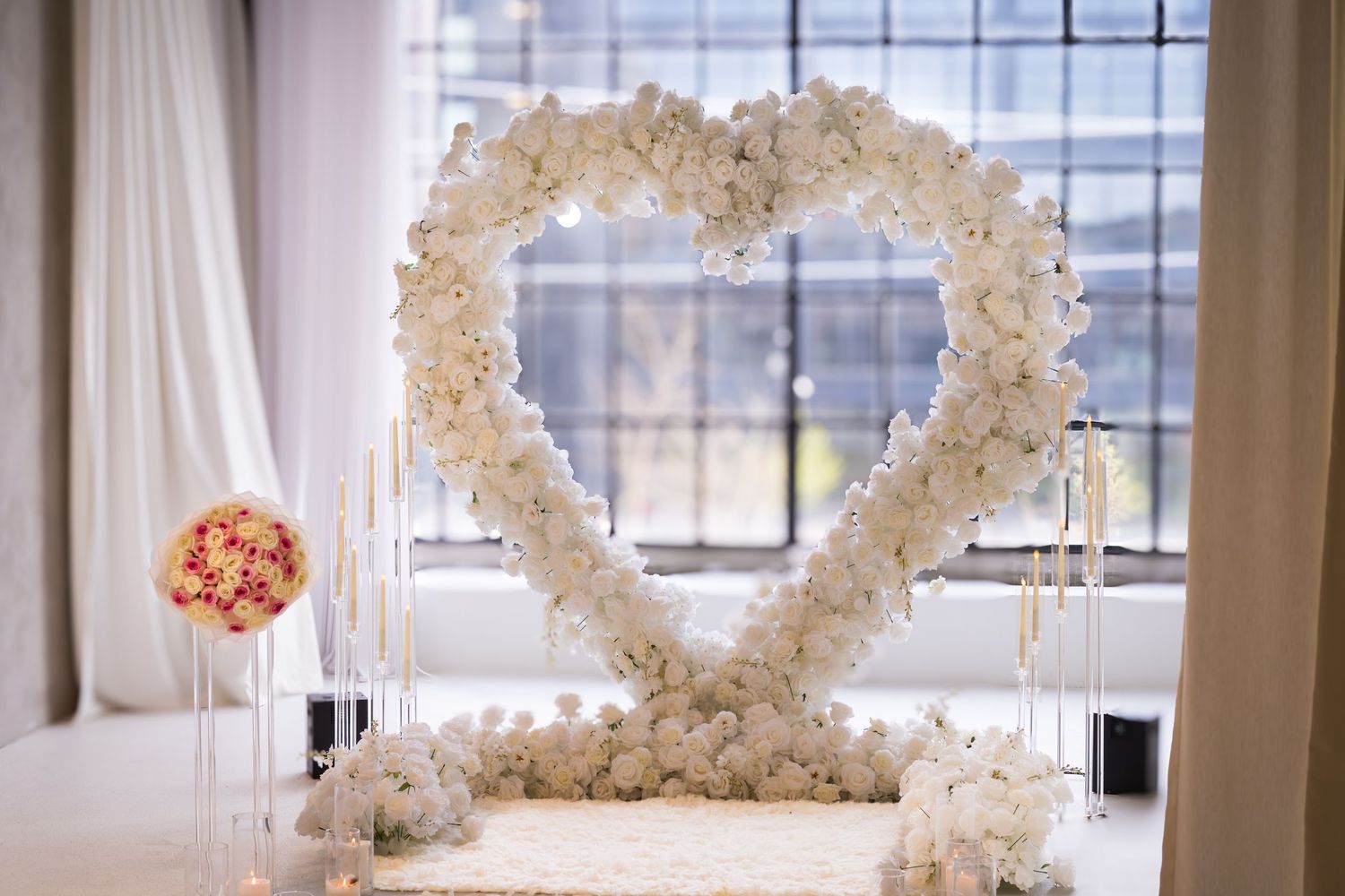 Elegant white floral heart-shaped arch with candles and roses in a bright, romantic proposal venue.