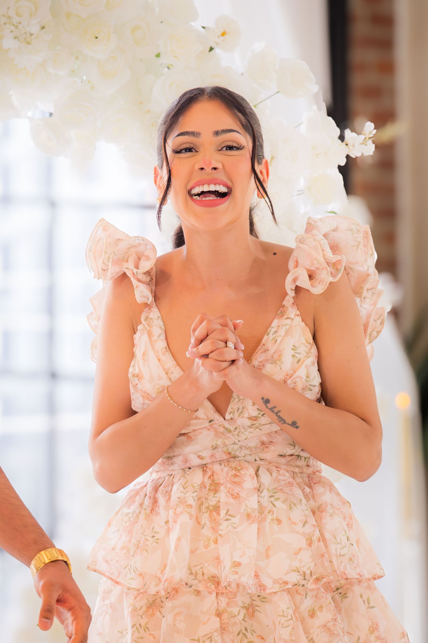 Joyful woman in floral ruffled dress laughing with clasped hands before white flower backdrop.