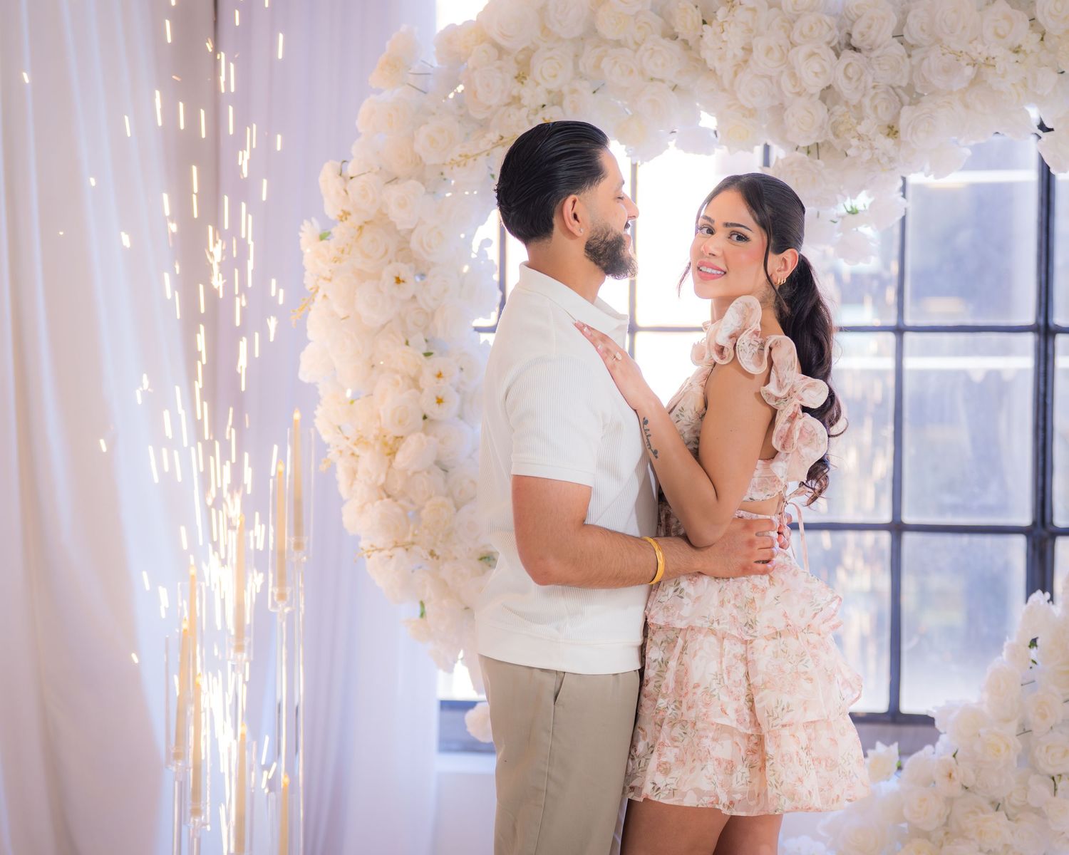 Couple embracing romantically inside a heart-shaped white floral arch with sparkling cold fireworks.