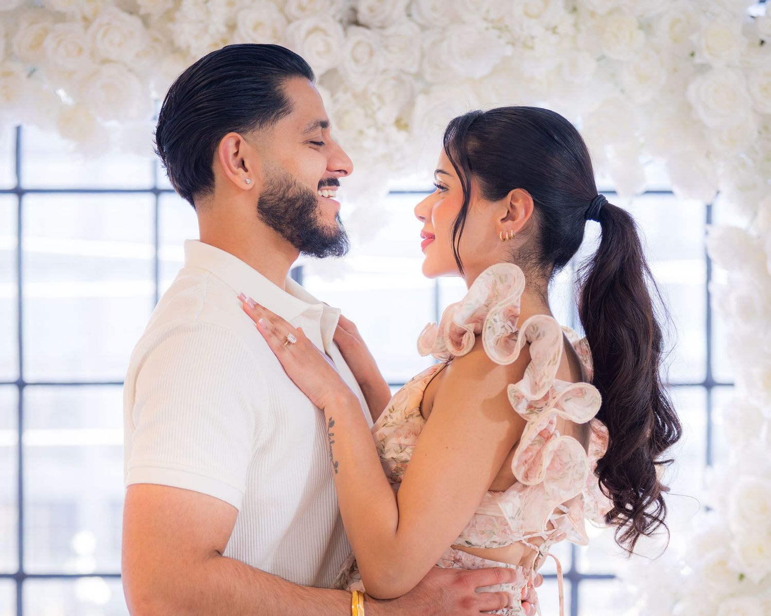 Smiling couple embracing and gazing at each other in front of a lush white floral wall.