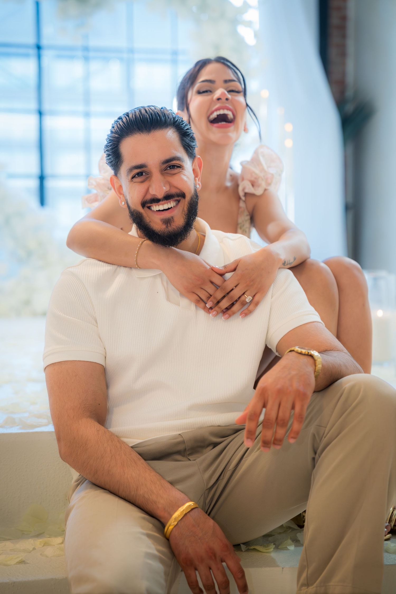 Happy couple laughing and embracing during romantic engagement photoshoot with white floral backdrop.