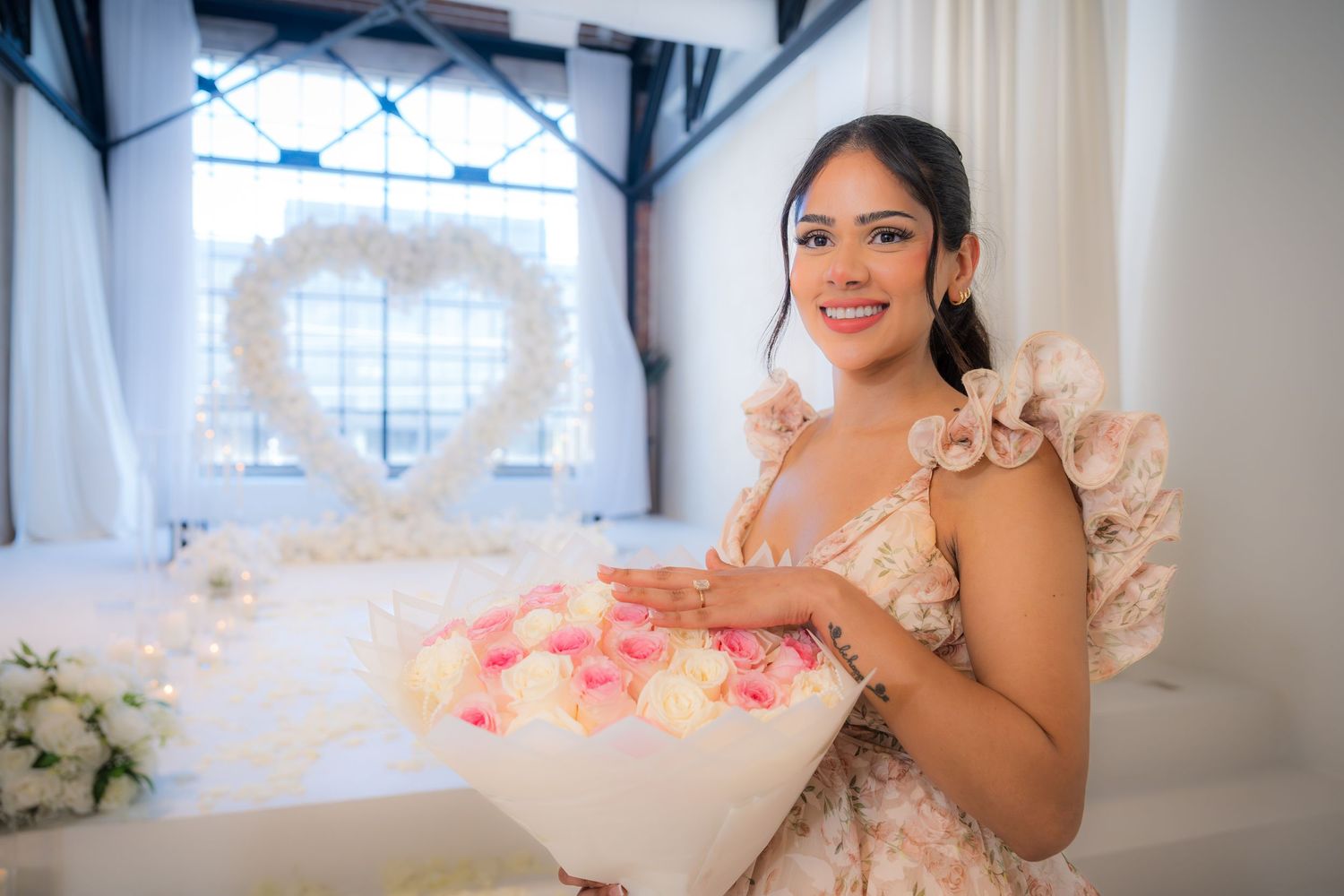 Young woman in floral dress holding bouquet of pink and white roses with heart-shaped floral backdrop.
