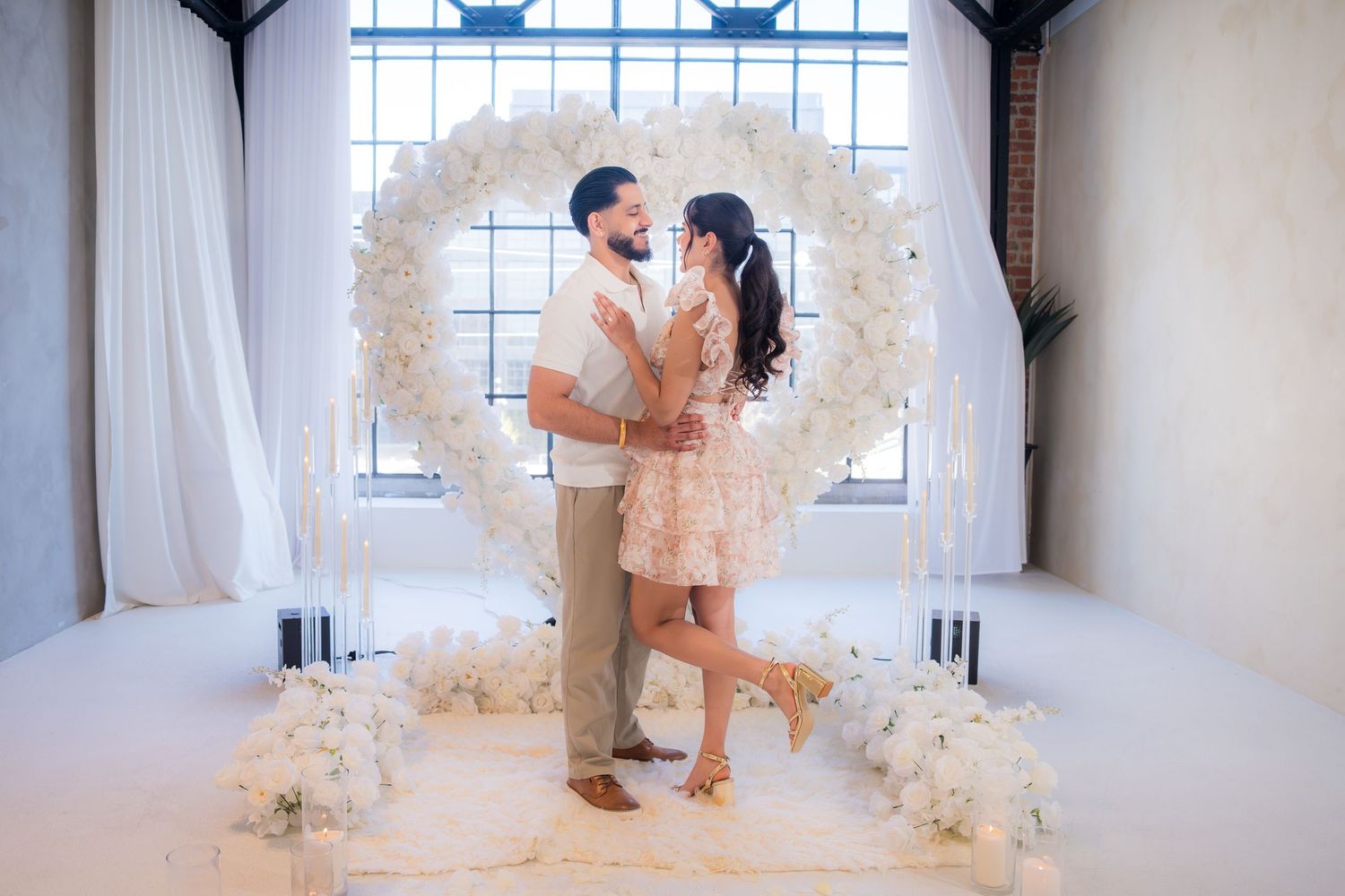 Couple standing face to face before large white heart floral arch with candles.