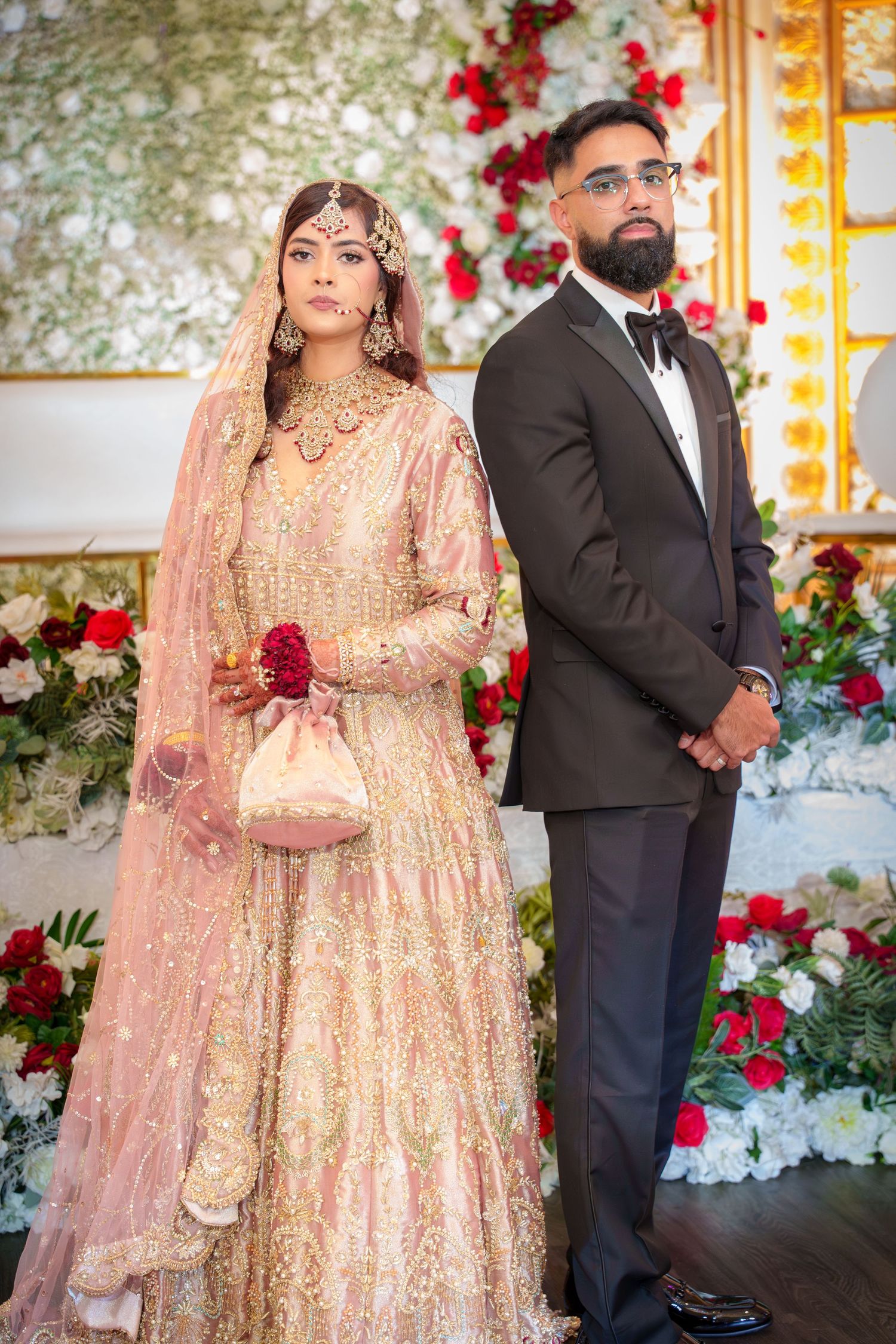 Traditional South Asian wedding couple in peach and black attire pose against floral wall backdrop.