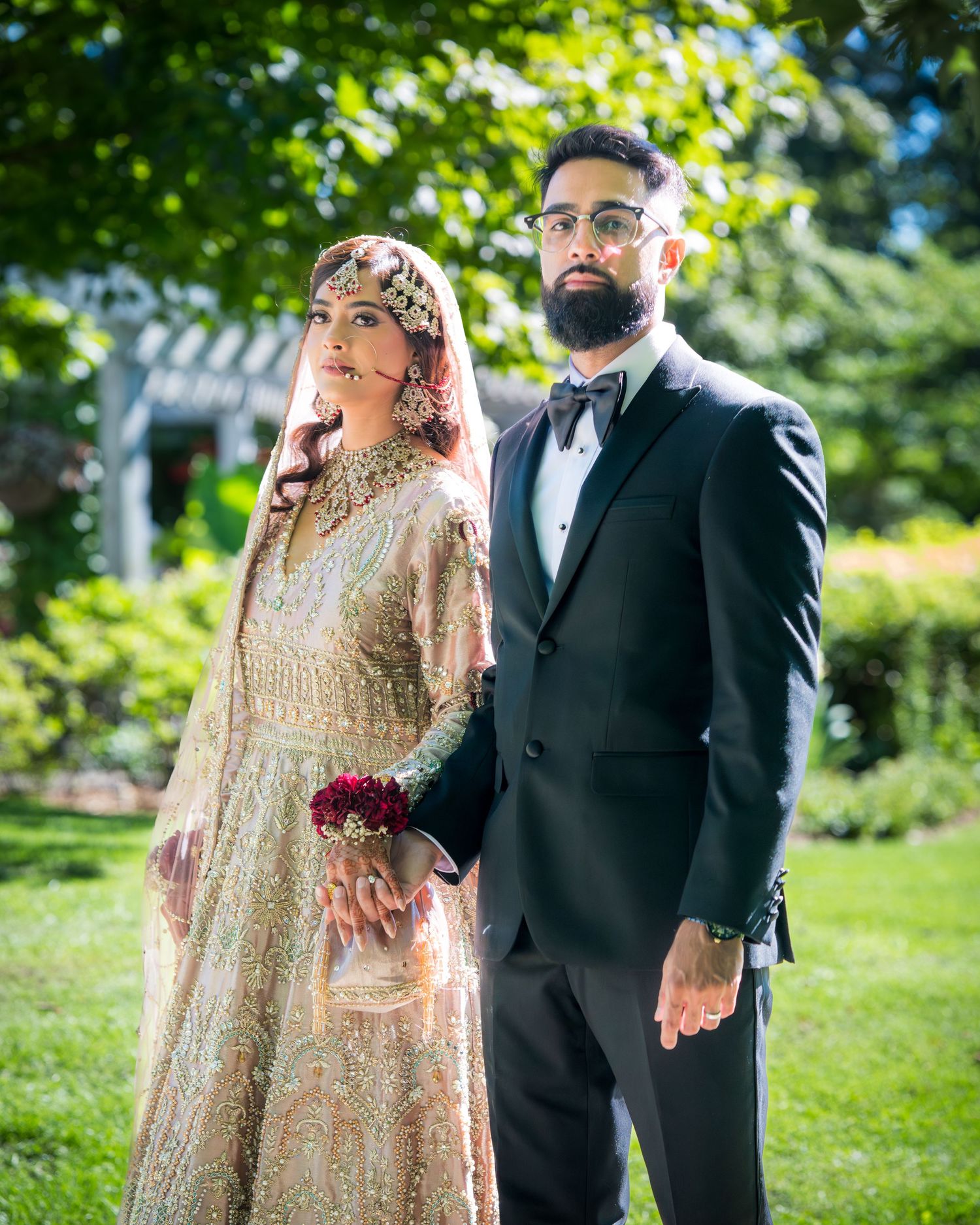 A couple in traditional South Asian wedding attire pose together outdoors in a garden setting.