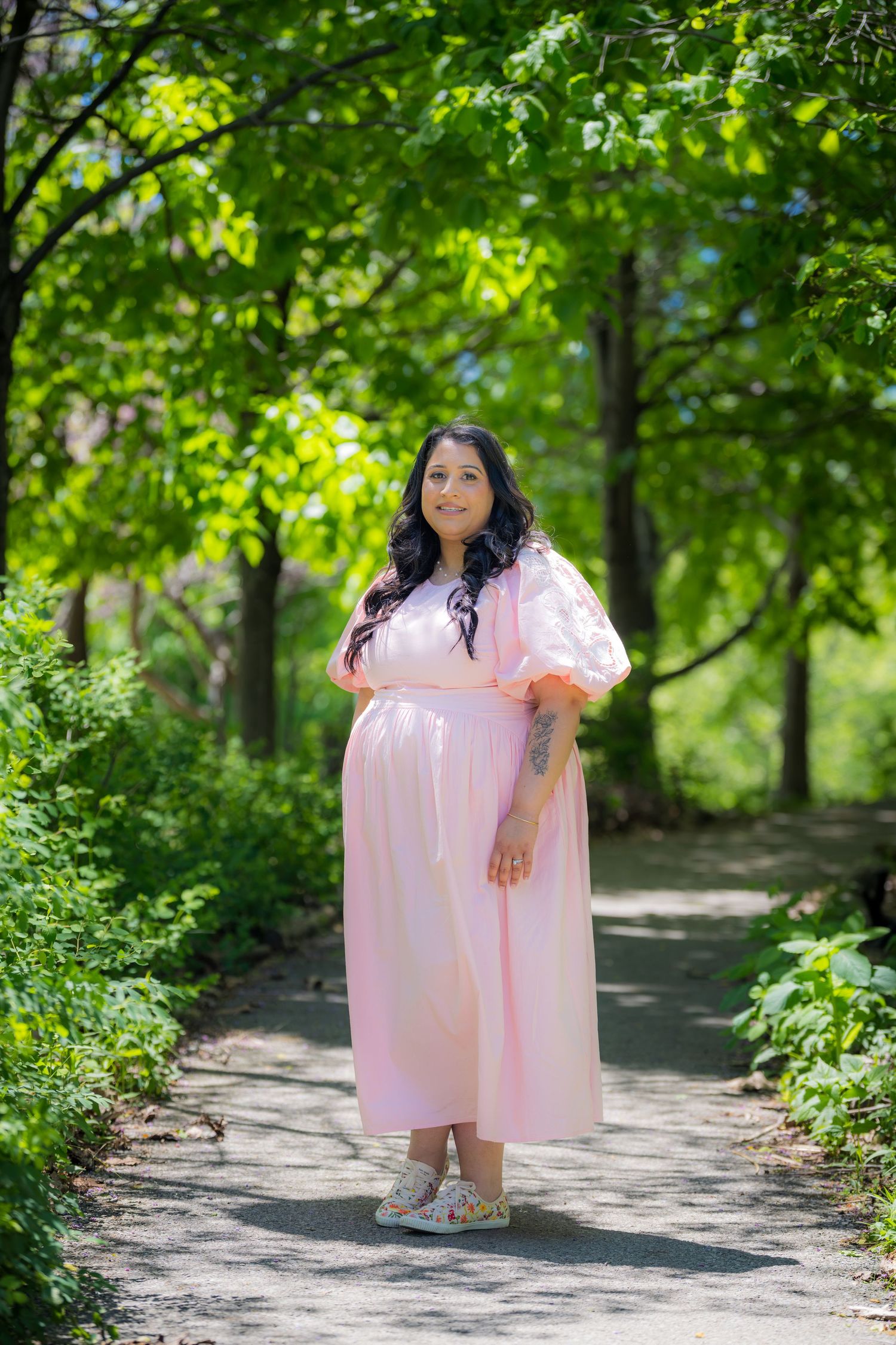 Pregnant woman in light pink puff sleeve dress walks along tree-lined garden path.