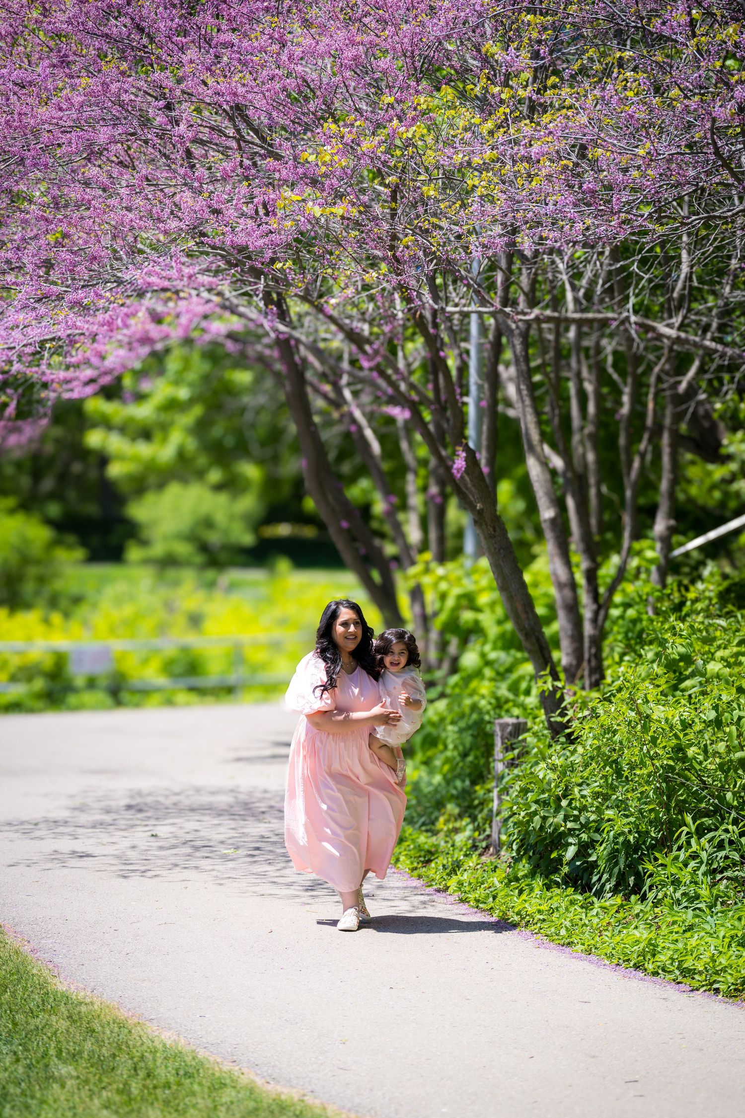 Pregnant woman in pink dress carrying toddler while walking under blooming purple redbud trees.