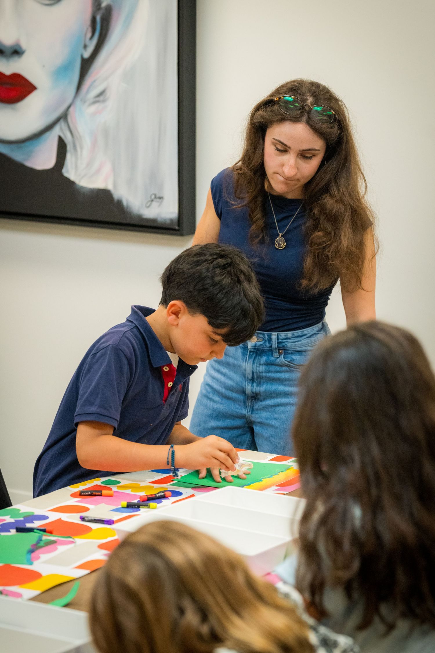 Female art teacher assists young boy with colorful paper collage project in bright art classroom.