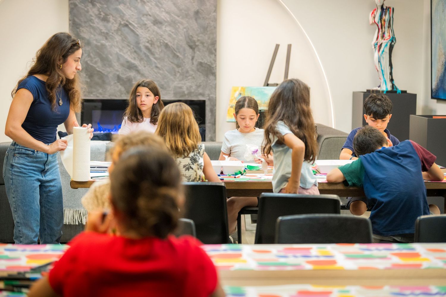 Children engaged in an art class activity at a table, guided by a teacher in a bright modern studio.