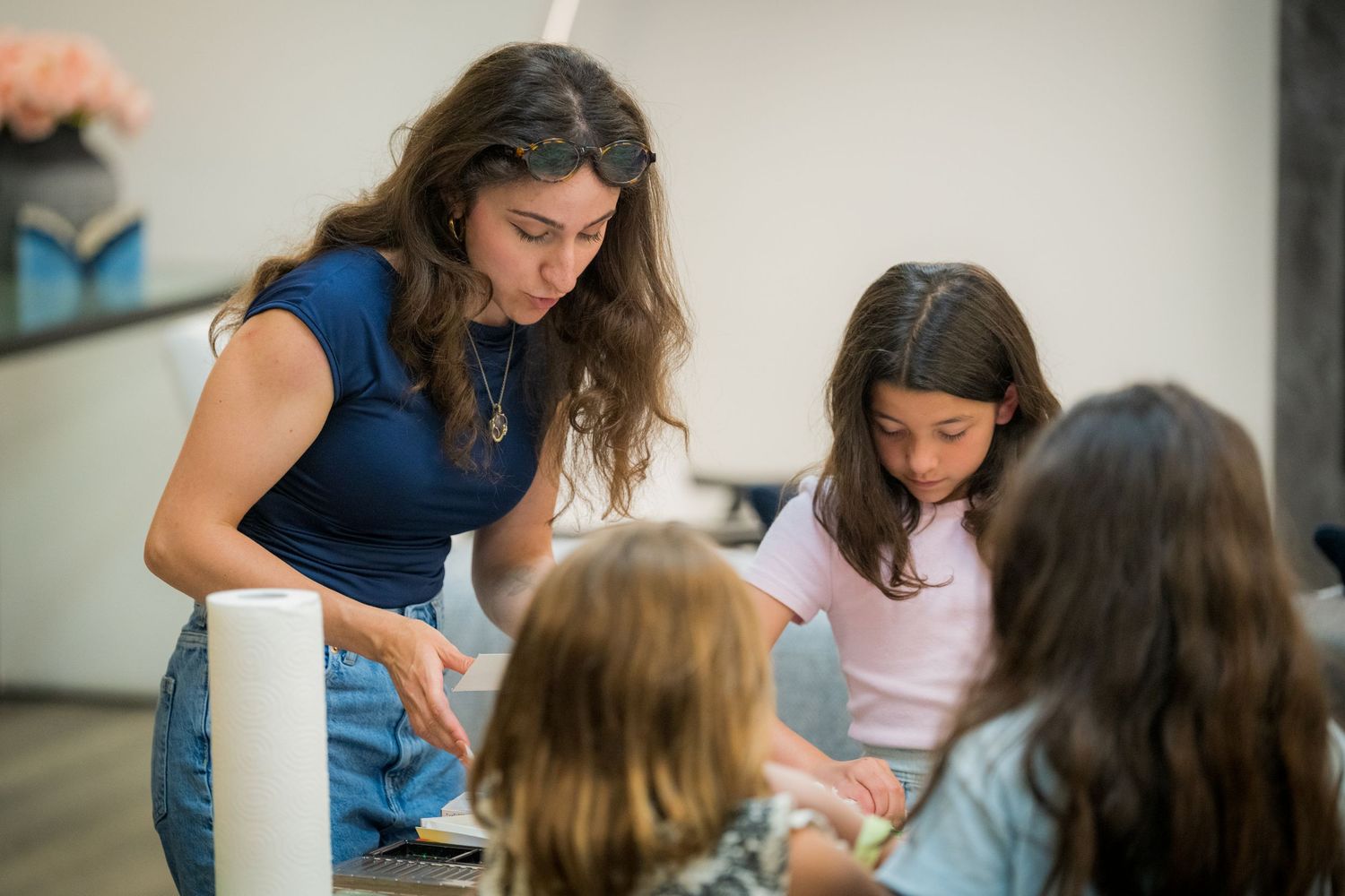 A teacher engages with three young girls during a hands-on learning activity at a bright, modern classroom table.