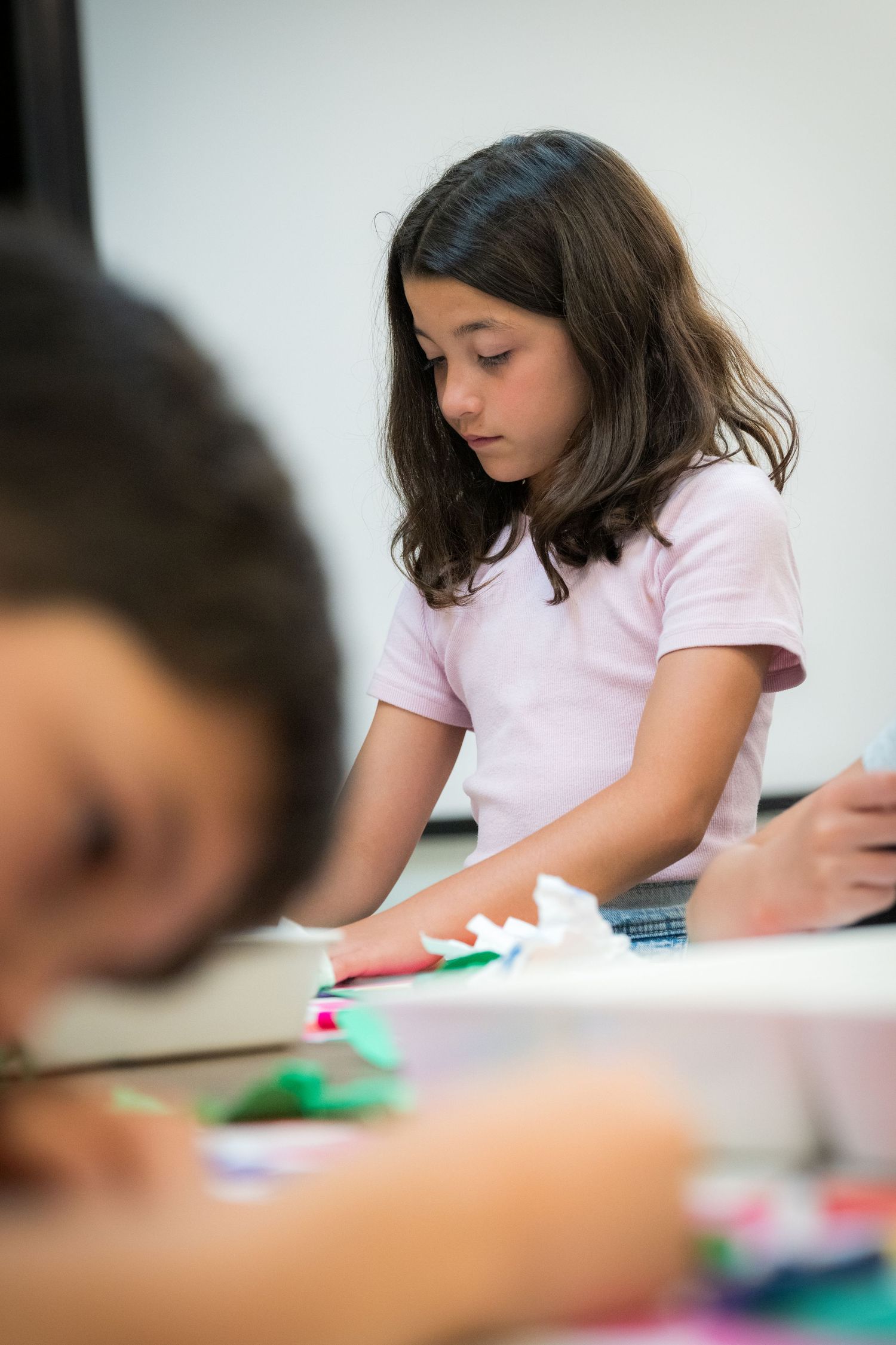 Young girl in pink shirt focuses on colorful paper craft project at art class table.