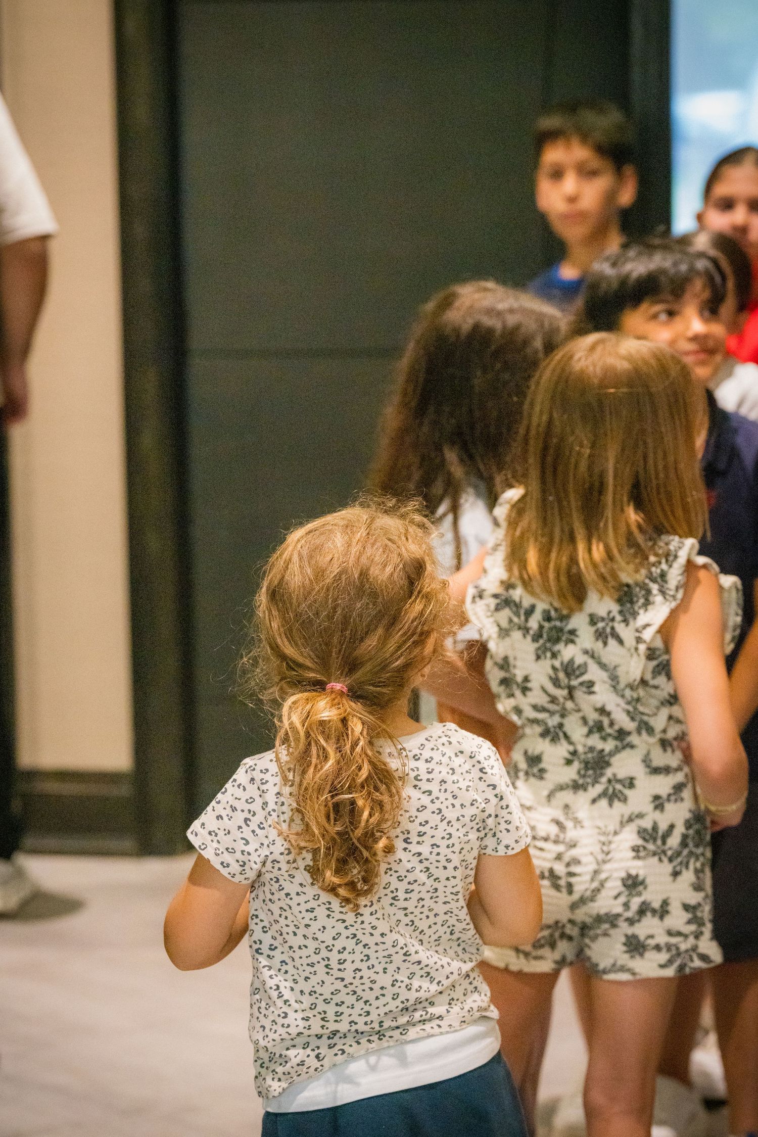 Group of children standing together indoors, viewed from behind during a school activity.