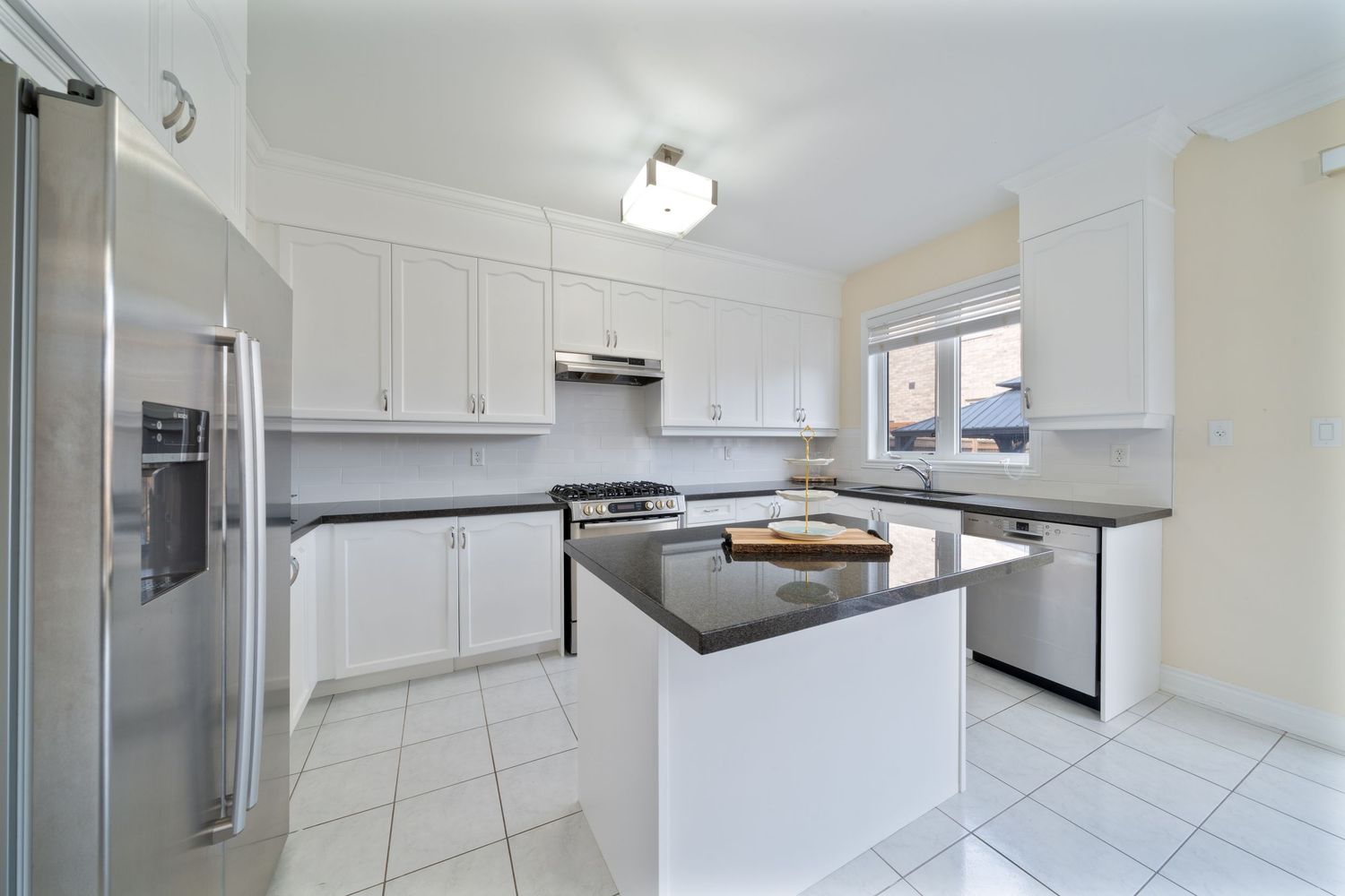 Modern white kitchen with dark granite countertops, center island, stainless steel appliances and tiled flooring.