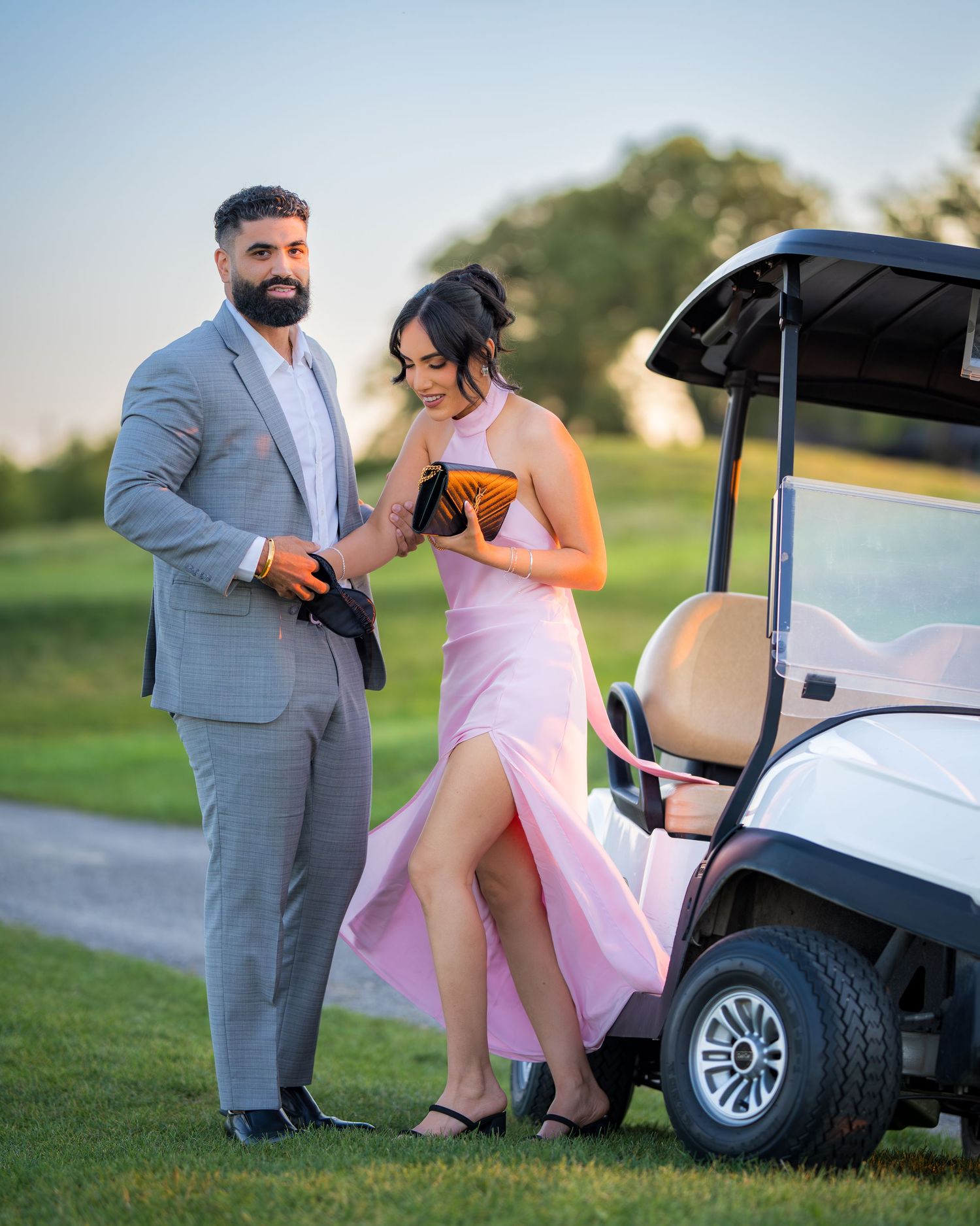 A couple stands near a golf cart on a grassy hill at sunset.